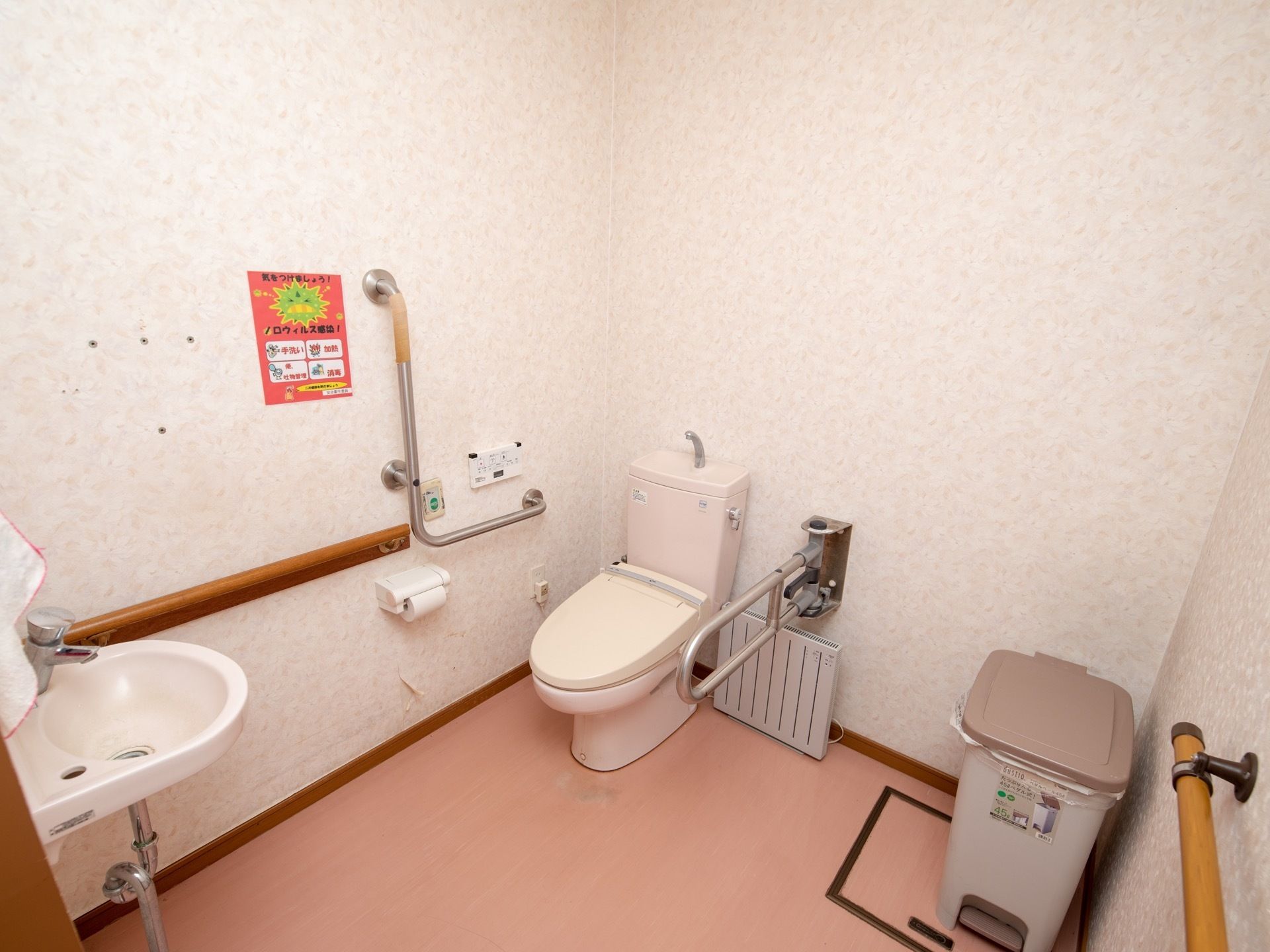 白色の壁、ピンク色の床に施工されたトイレである。壁にはステンレス製の手すりが設置されている。ゴミ箱もある。