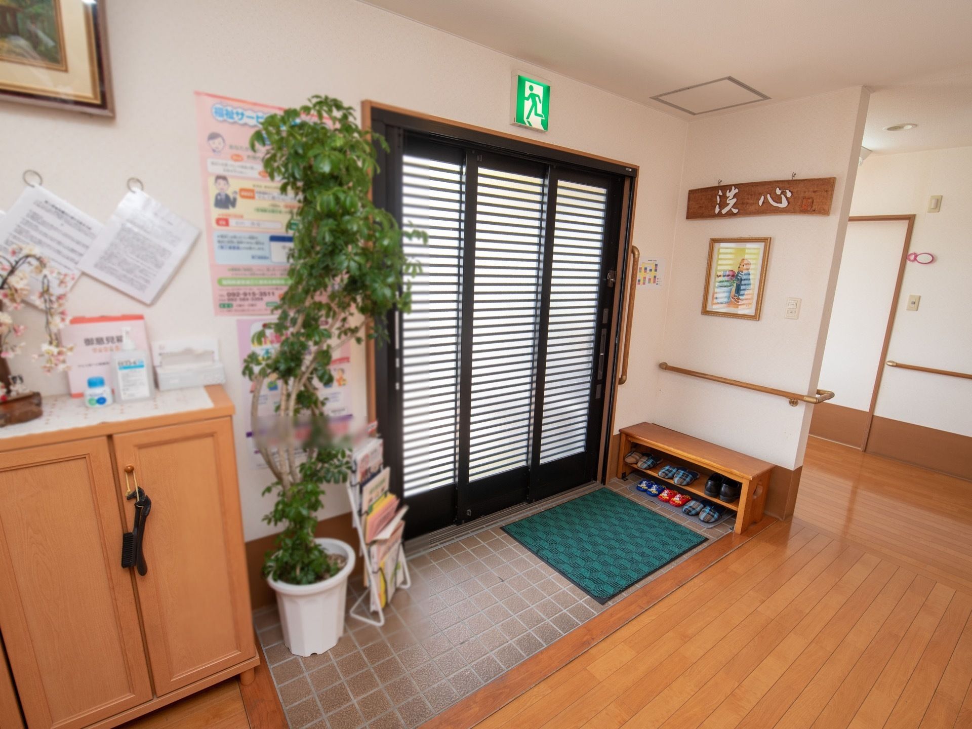 目隠しの枠が設置された、３枚連動扉が備えつけられている。上部に非常口誘導灯がある。背丈の高い、観葉植物が置かれている。