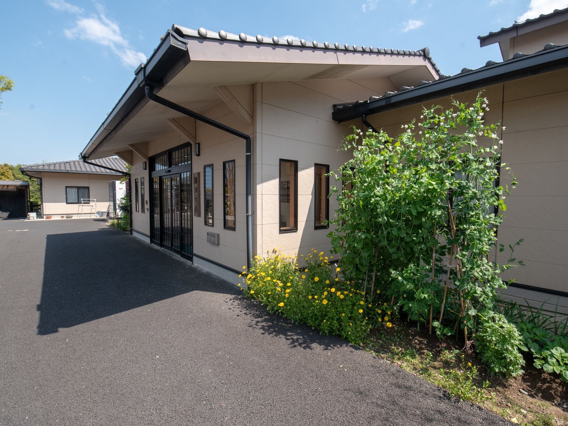 クリーム色の外観である。１階建てで、その前にアスファルトの敷地が広がっている。緑が生い茂った、家庭菜園も設置されている。