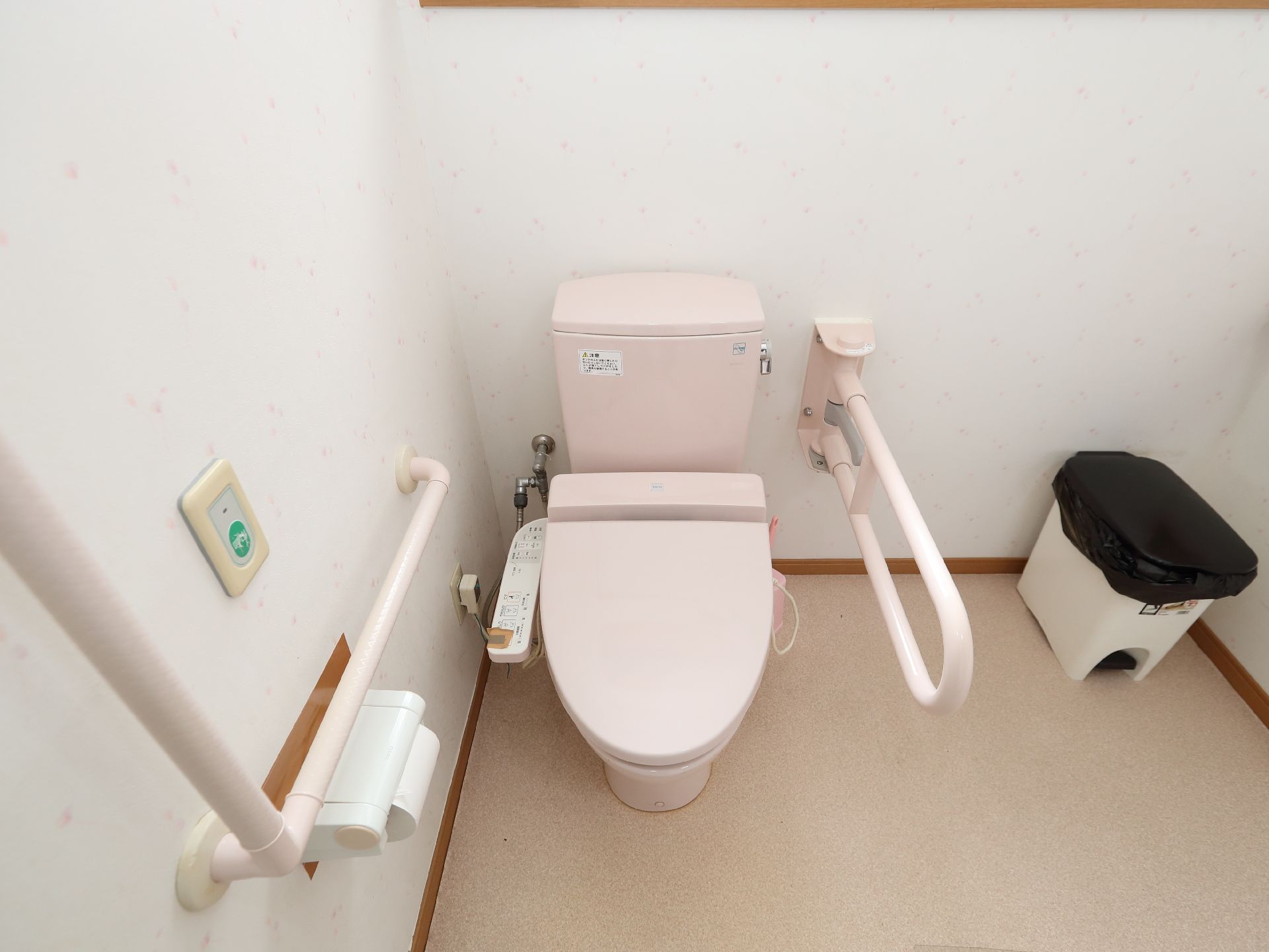 白い壁の部屋の一角に設けられた手すりに囲まれたピンク色の洋式便器