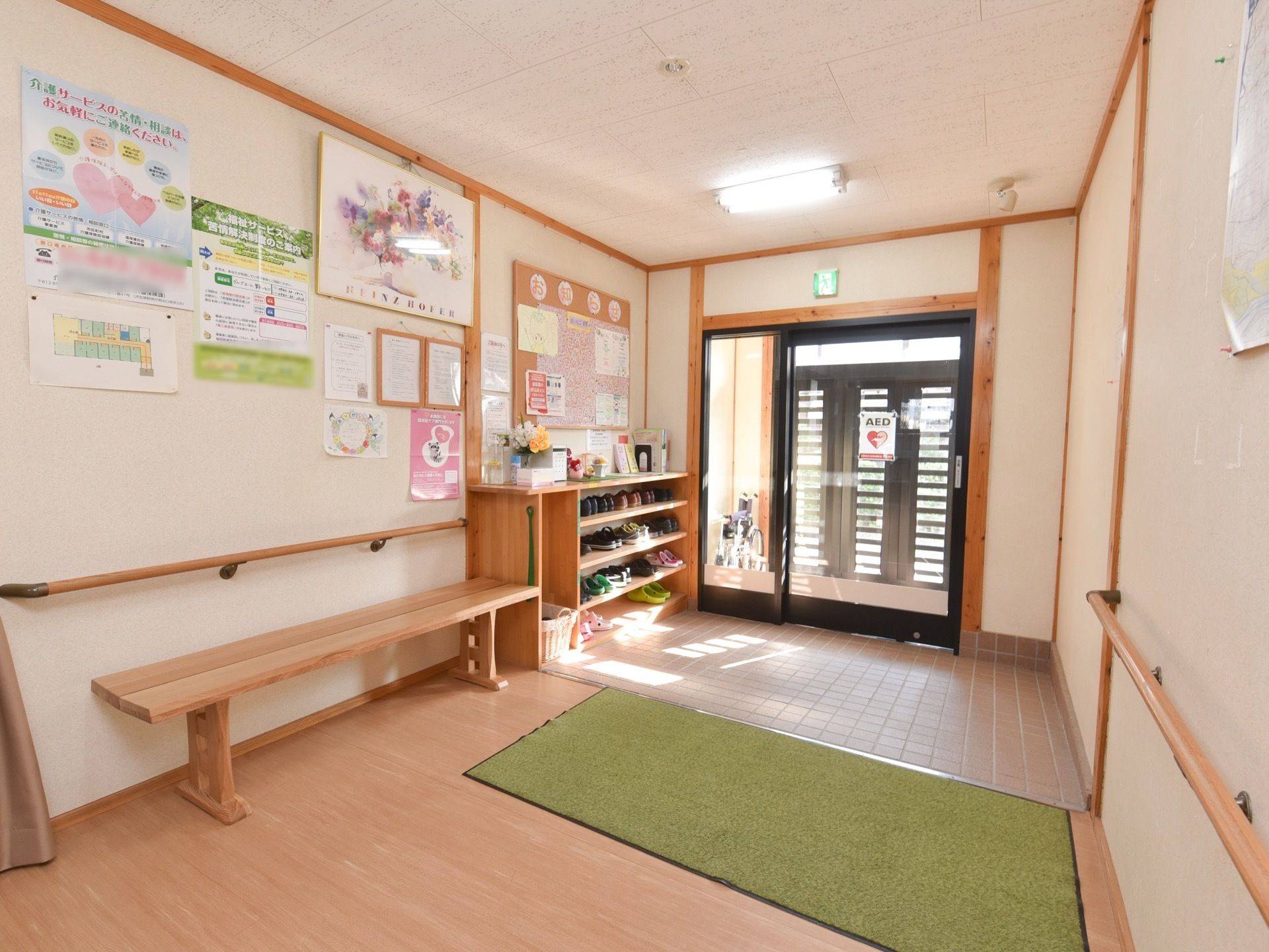 黒い扉とガラス張りの自動ドアの二重扉がついている玄関。床はタイルになっており、玄関マットが敷かれている。下駄箱とベンチが置かれている。