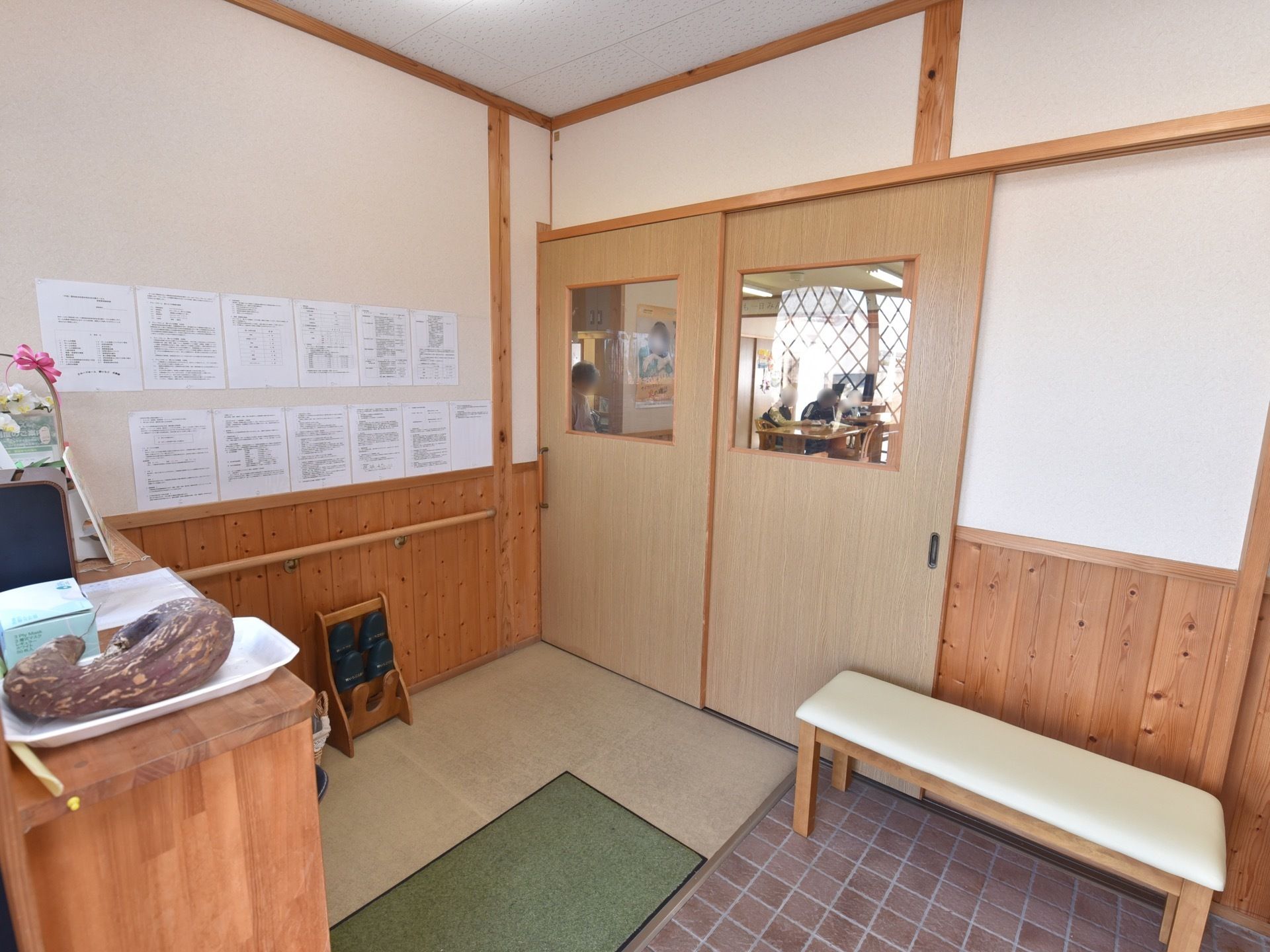 玄関に通じる扉がある。一部がガラス張りになっており、中の様子が伺える。壁には張り紙が貼られ、ベンチが置かれている。