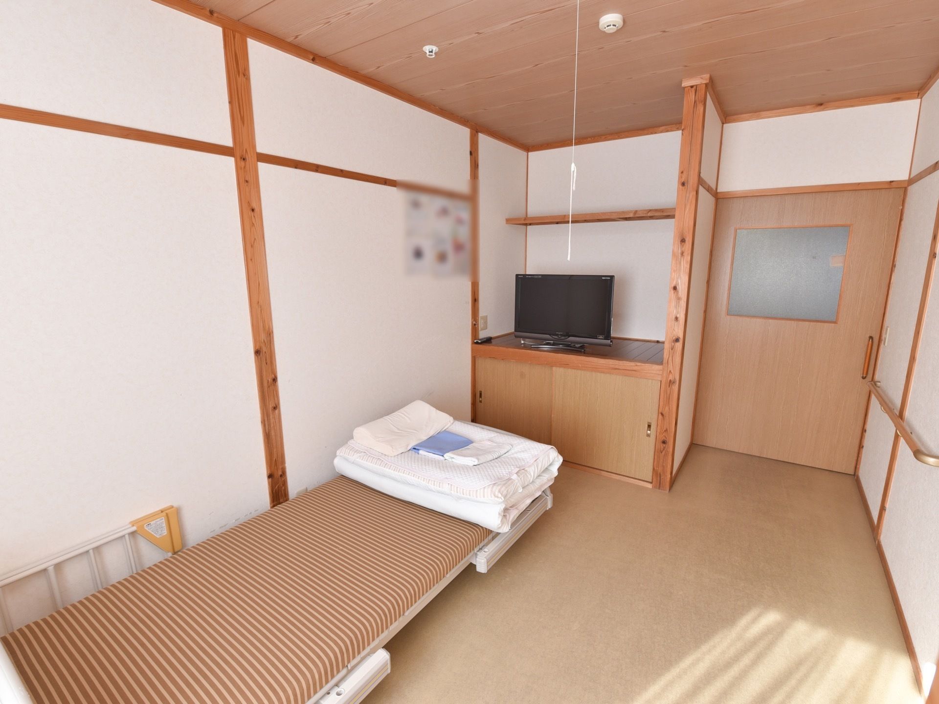 木目調の木の柱がある入居者様の居室。介護用ベッドが置かれている。壁の一部が大きな棚になっており、テレビが置かれている。