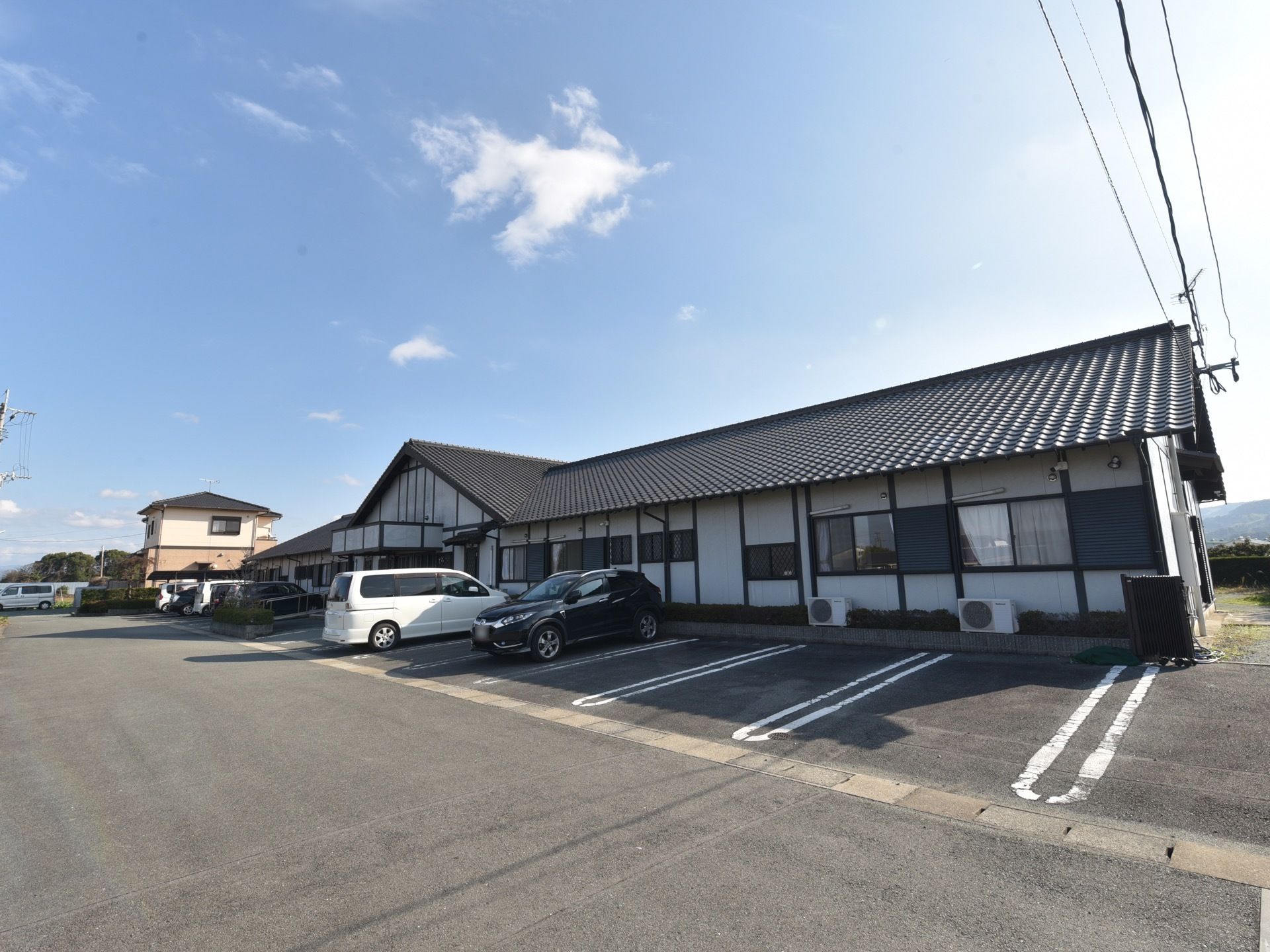 黒い大きな瓦屋根のあるの本家屋風の建物。敷地内は広く、建物の前面部分が駐車場になっていて、車が数台停まっている。