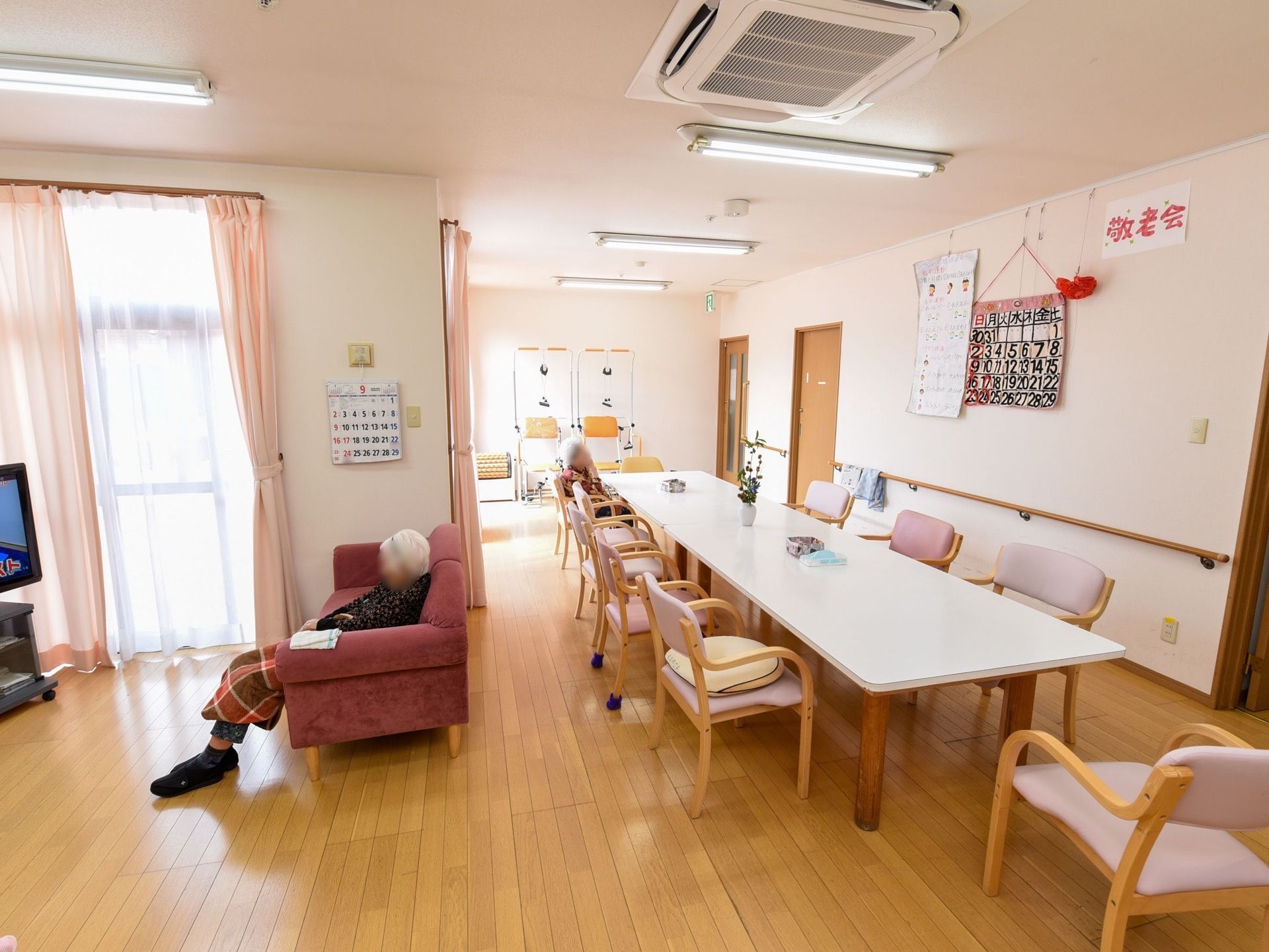 明るく広いリビングダイニングである。10脚の椅子がセットされたダイニングテーブルが置かれている。窓際には、ソファとテレビが設置されている。