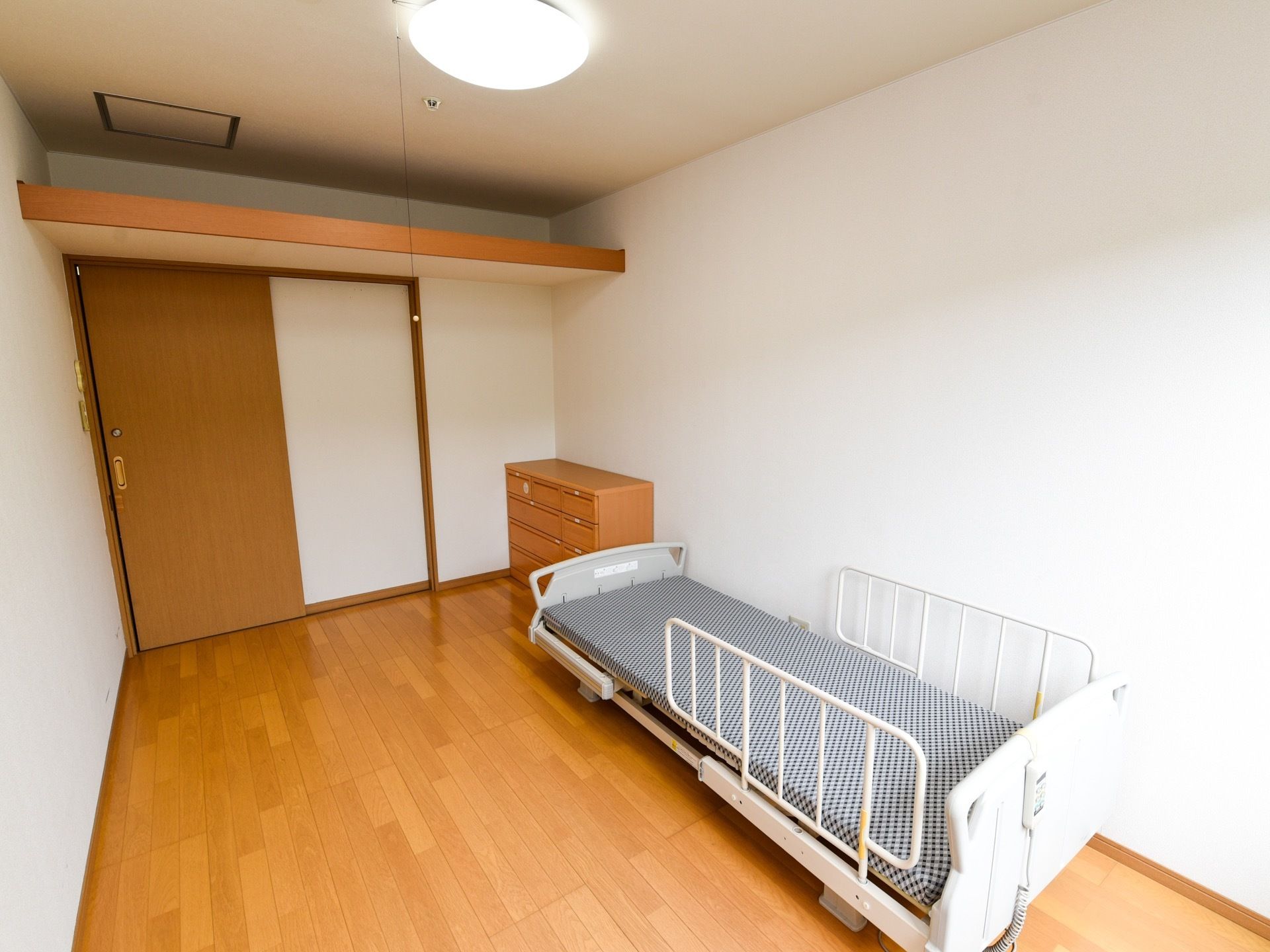 広めの間取りの居室である。介護ベッドやタンス、照明器具が完備されている。大きな棚が扉の上に備えつけられている。