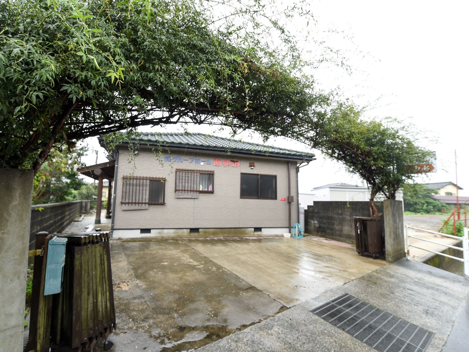 施設のグレー色の外壁には、紫色とピンク色で施設名が書かれている。建物の前には広いスペースがあり、車を駐車することができる。