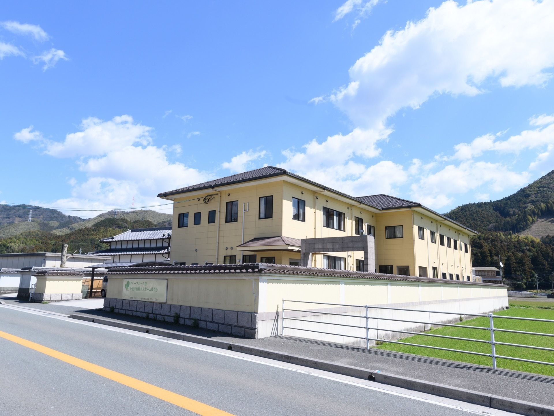 クリーム色の2階建ての建物になっている。建物の前には道路が通っており、歩道や畑側にガードレールが設置されている。