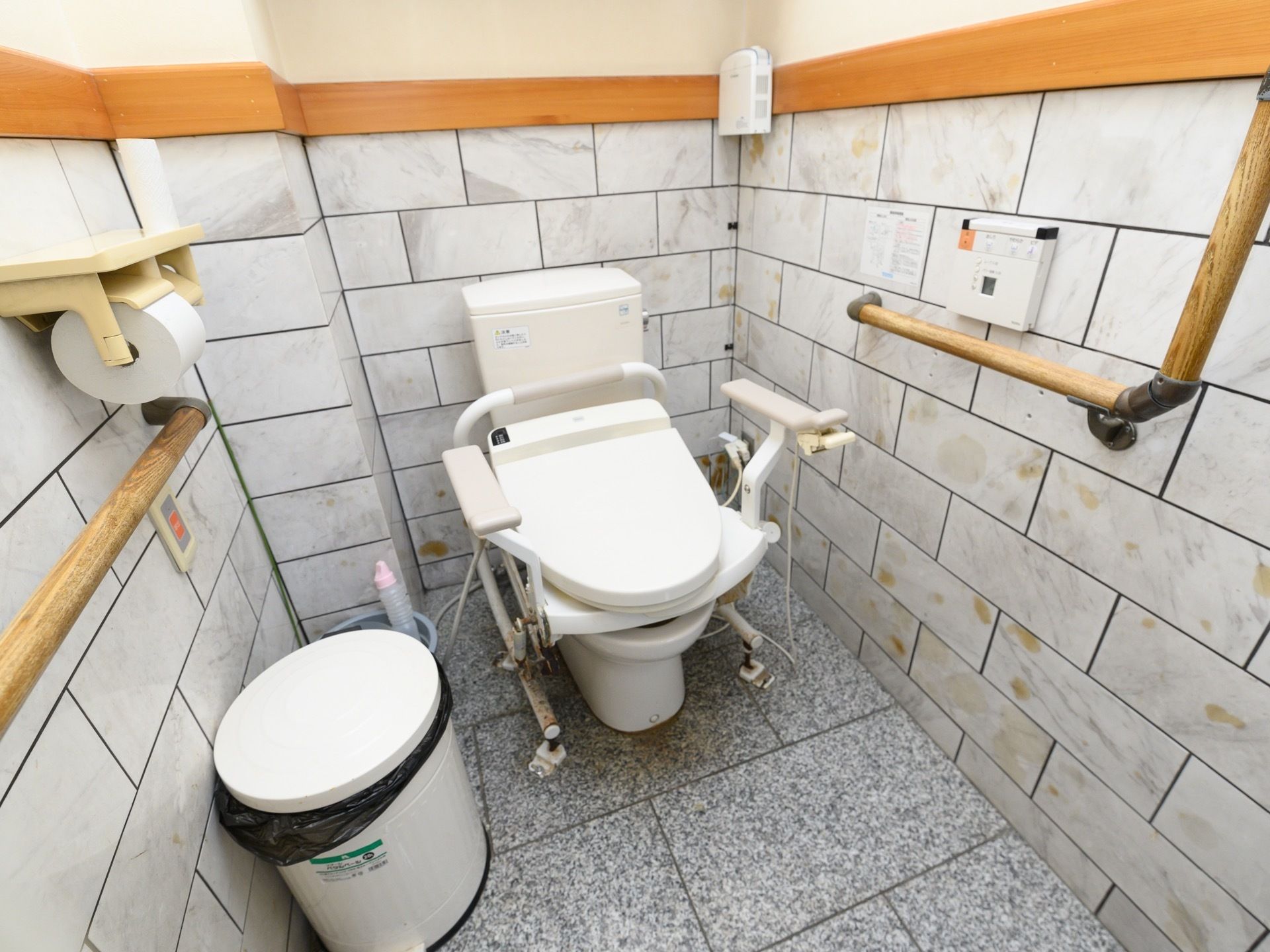 大理石調のグレーの床に、白い温水洗浄便座が設置されている。壁には手すりやトイレットペーパーホルダー、呼び出しボタンが取り付けられている。