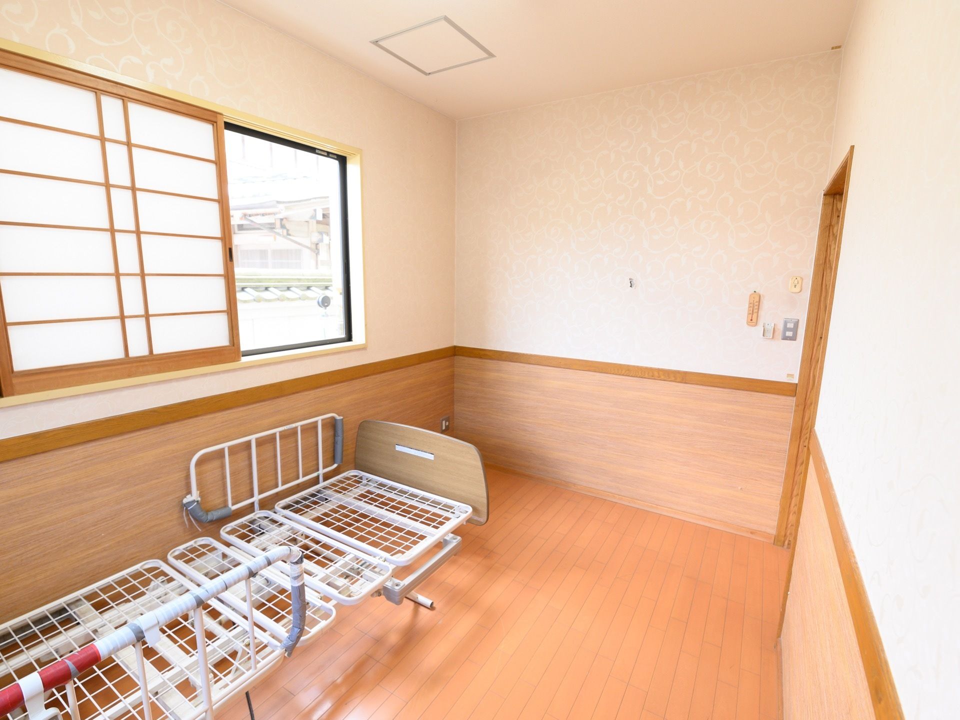 床と壁の下半分が木目調になっていて、壁際に介護ベッドが置かれている。腰高の窓には障子が取り付けられている。
