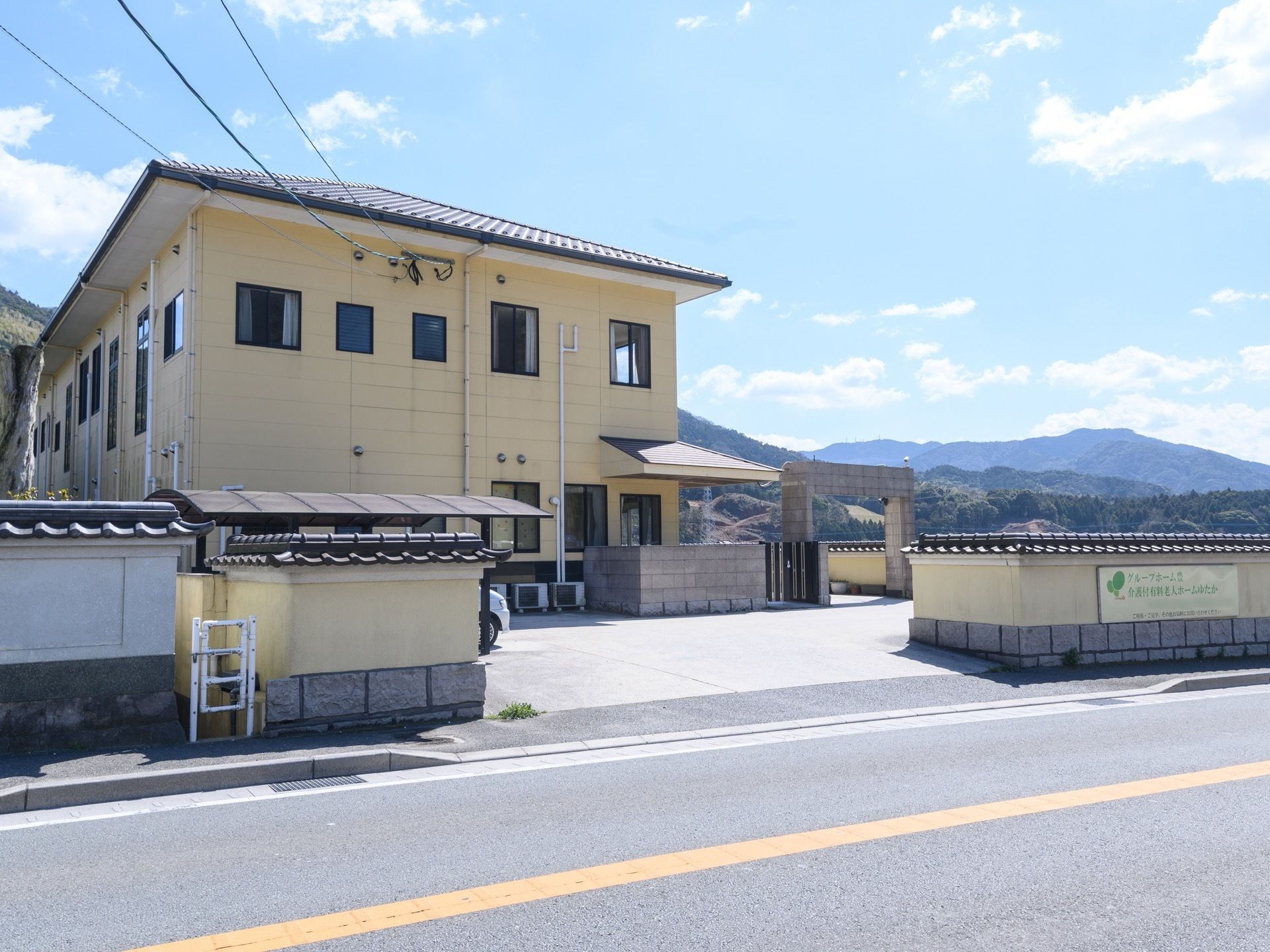 2階建ての建物になっていて、壁際にはエアコンの室外機が置かれている。道路からの出入り口近くの壁には、施設名の書かれた看板が取り付けられてる。