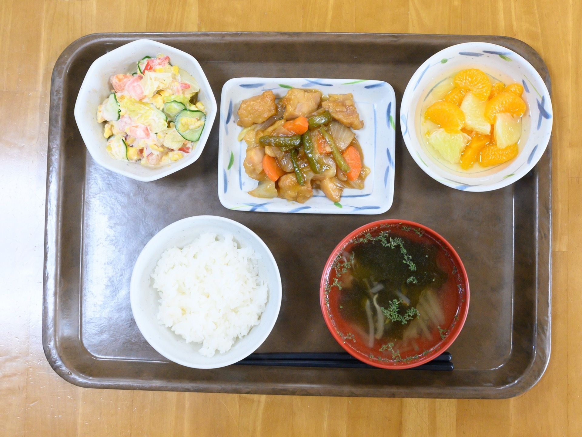 トレーの上に、ご飯と汁物、中央にメインおかず、その両側に小鉢とフルーツを添えた食事ある。栄養バランスと彩りにも配慮されたメニューである。