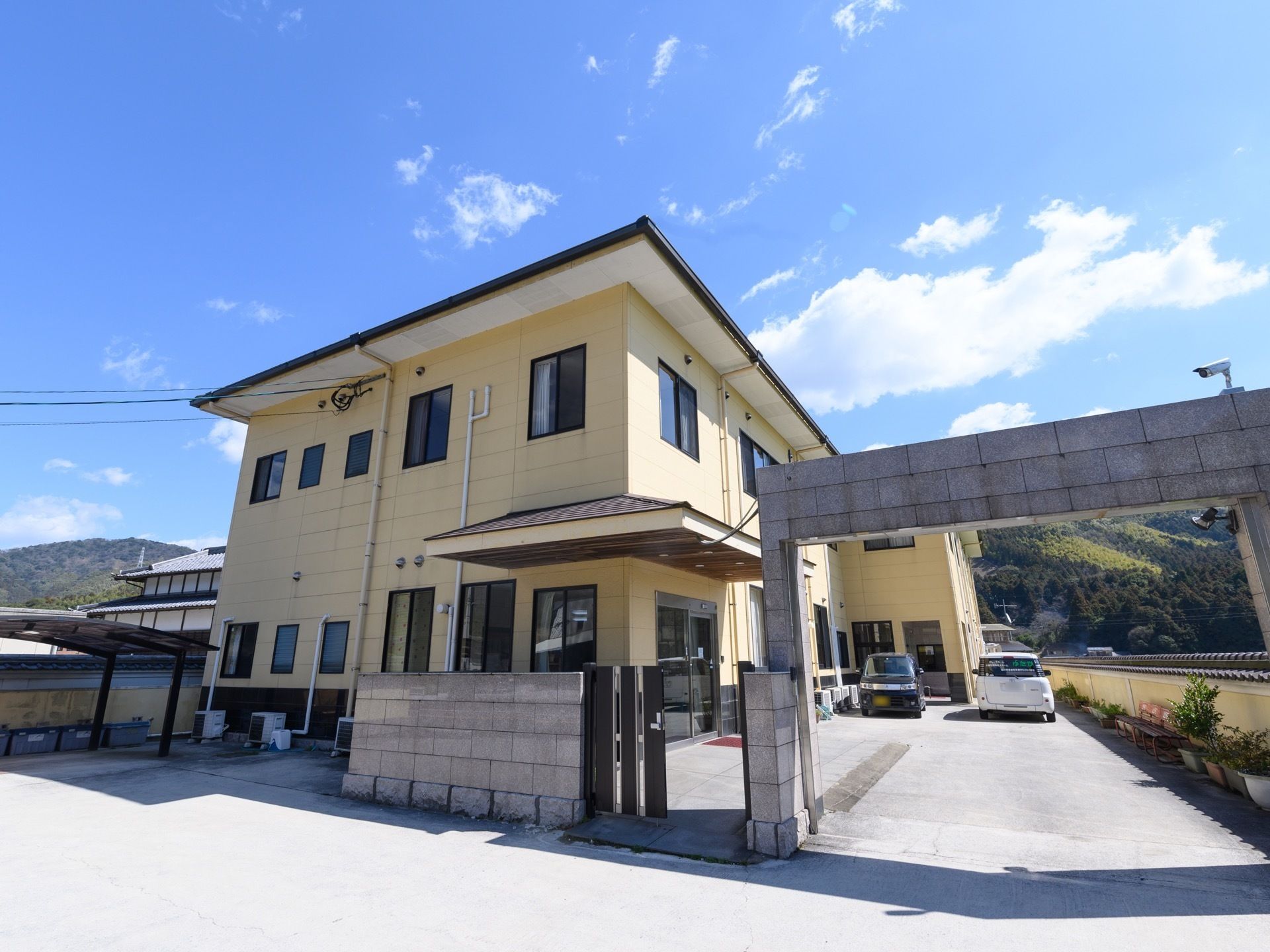 黄色い外壁に屋根は茶色で塗られている。カーポートが設置されており、その下にはプラスチックのボックスが複数置いてある。