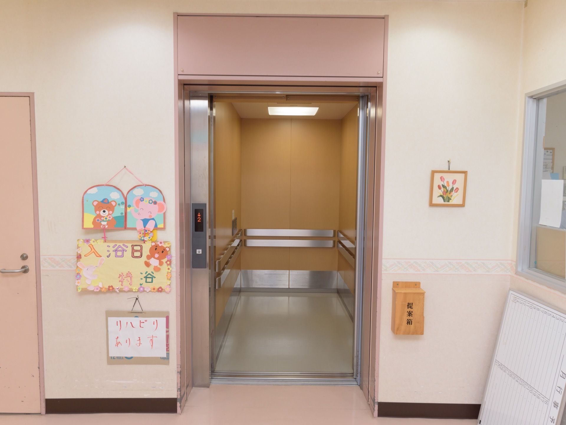 エレベーターが設置されている。乗降口の左側に、入浴日やリハビリ実施を知らせる表示がある。右側には提案箱が取り付けられている。