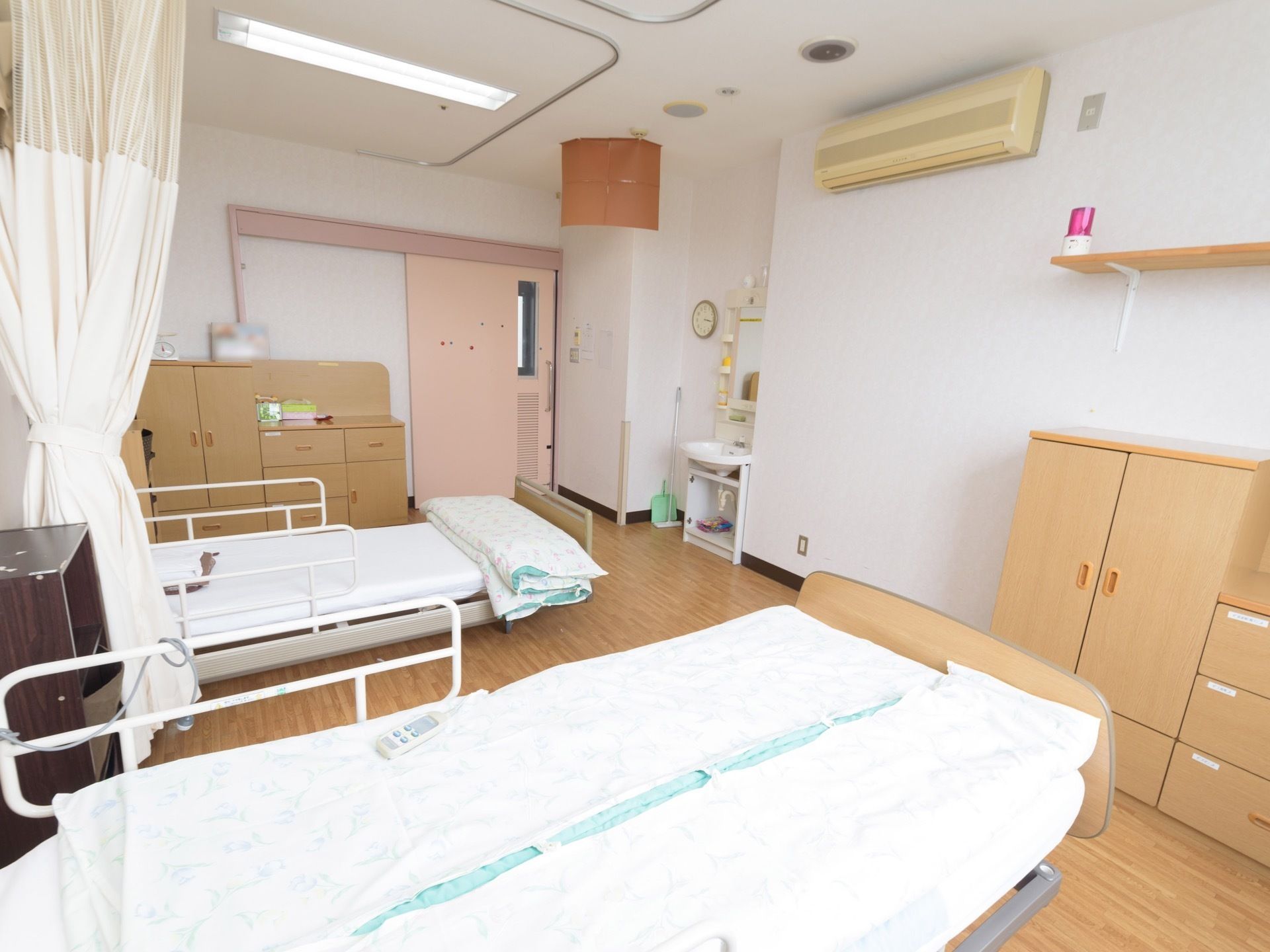 居室に、転落防止柵のついたベッドが２台置かれ、その間はカーテンで仕切れるようになっている。洗面台やエアコンが設置されている。