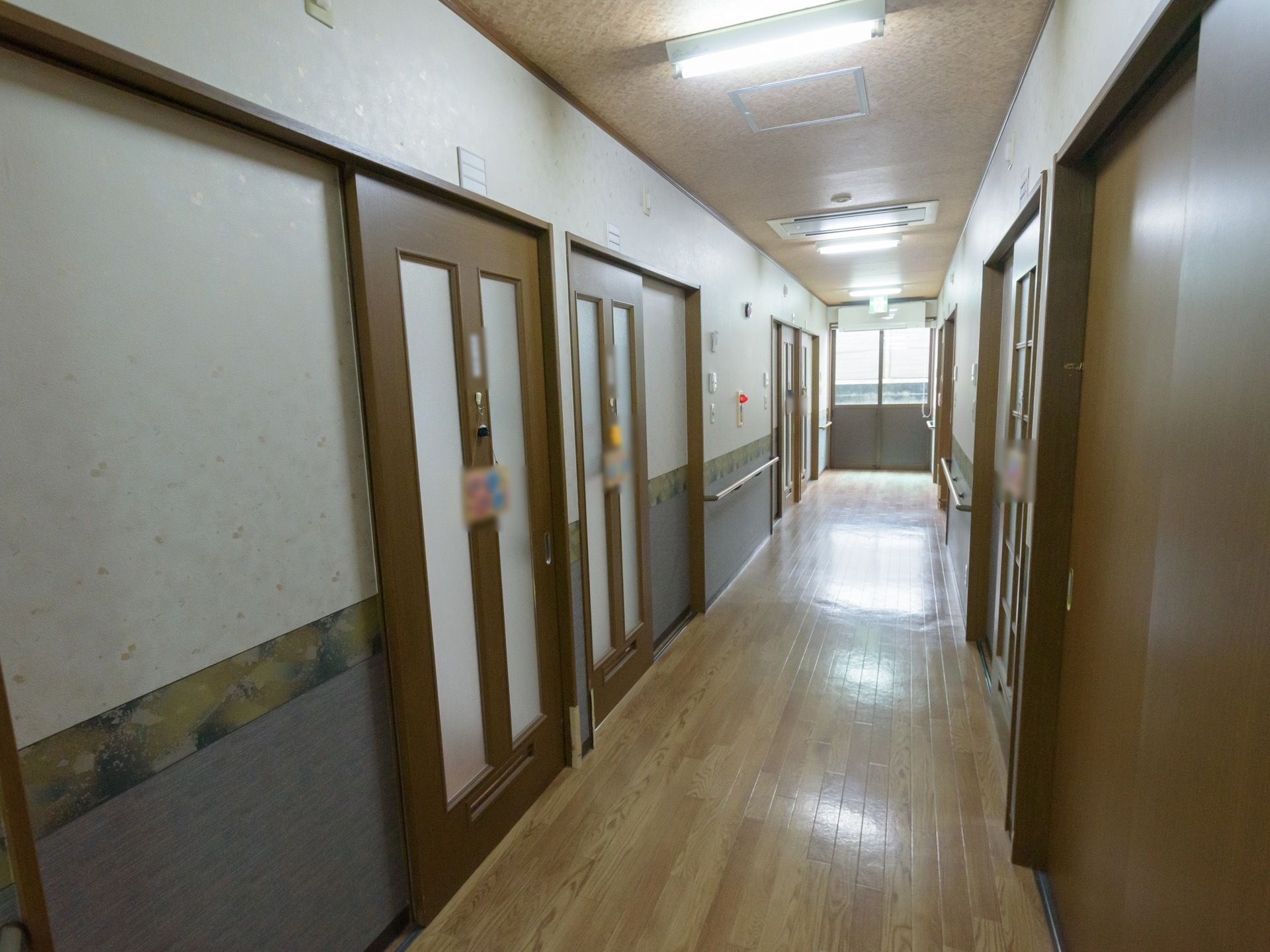 フローリング材の廊下がまっすぐ伸び、突き当りには窓がある。両側には居室へのドアが並び、ネームプレートがかけられている。