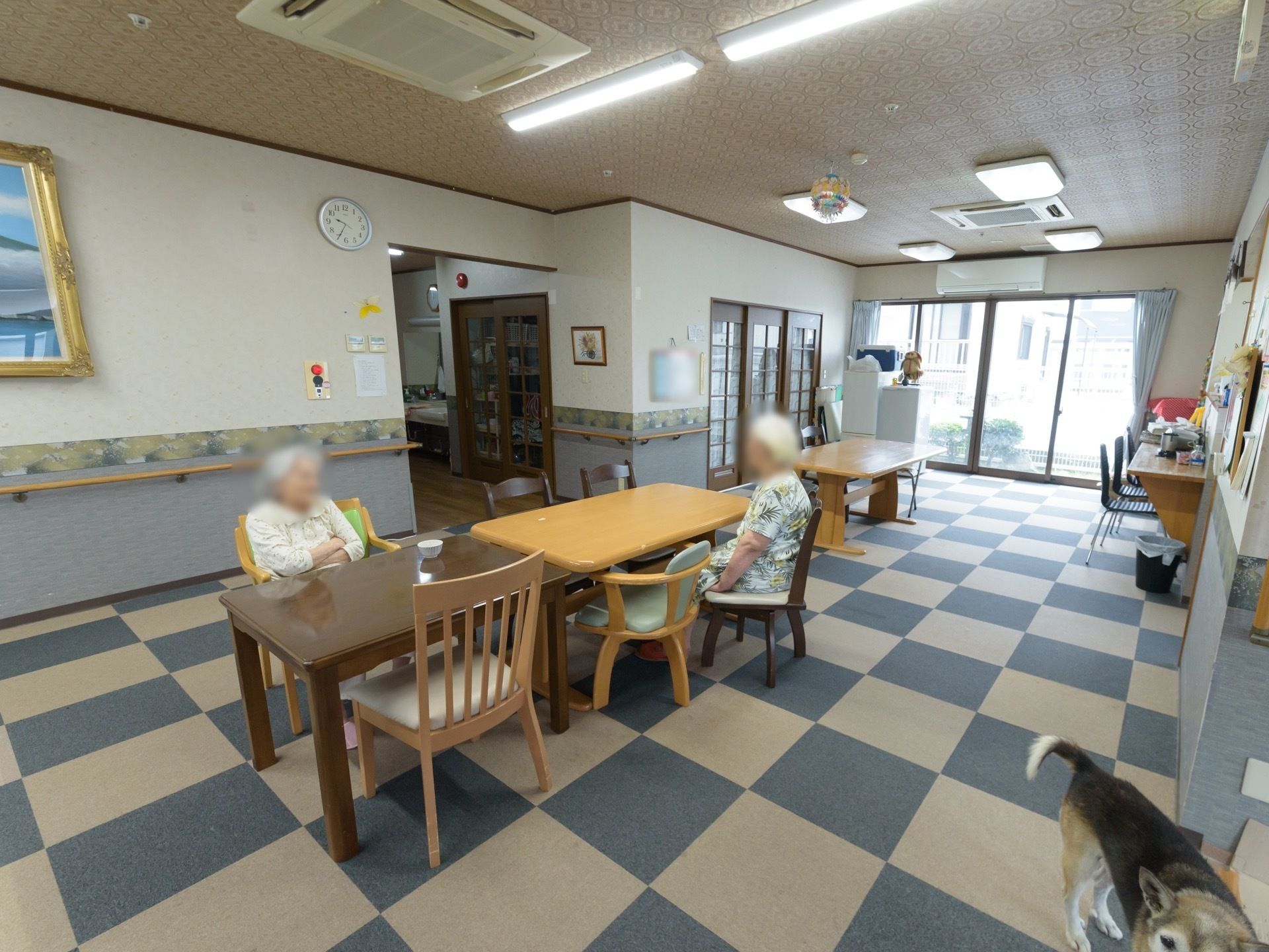 格子模様に絨毯が貼られた共有スペースには、テーブルがたくさん置かれている。その周囲で入居者様がくつろいでいる。