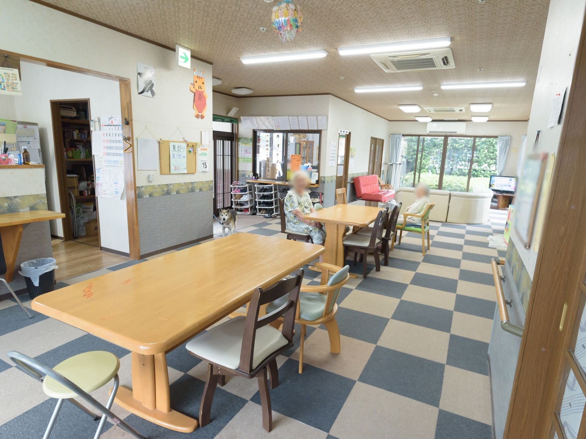 開放的な共有スペースでは入居者様が椅子に腰かけている。テーブルがいくつも並んでおり、奥にはテレビやソファーがある。