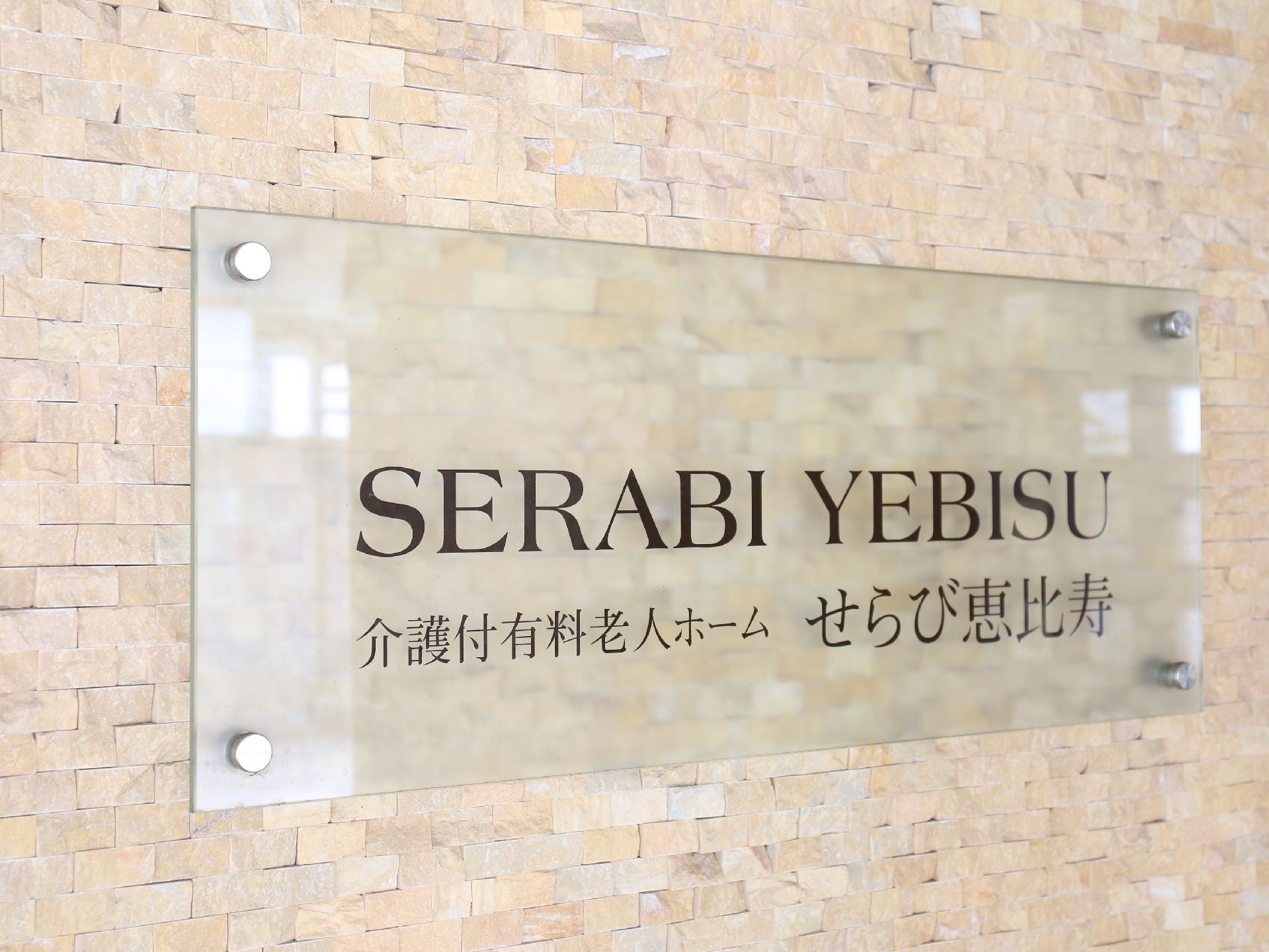 クリーム色のレンガ調の壁に、ガラス風のプレートの看板が掛けられている。看板には黒い文字で、施設名とロゴが書かれている。