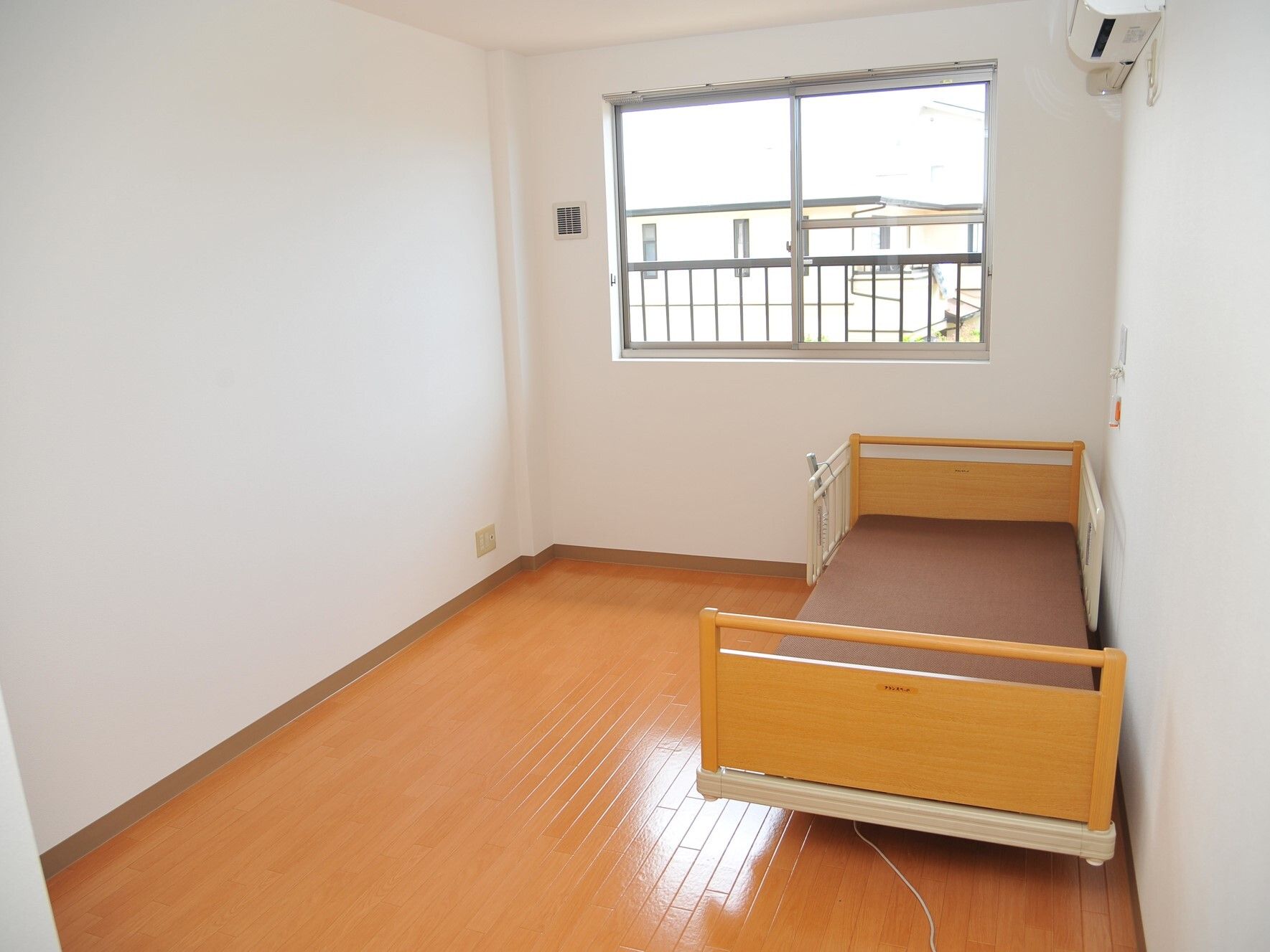 白い壁にフローリングのフロアと明るい雰囲気の部屋。介護用ベッドには寝具はなく、すぐそばに緊急時にもが付いている。