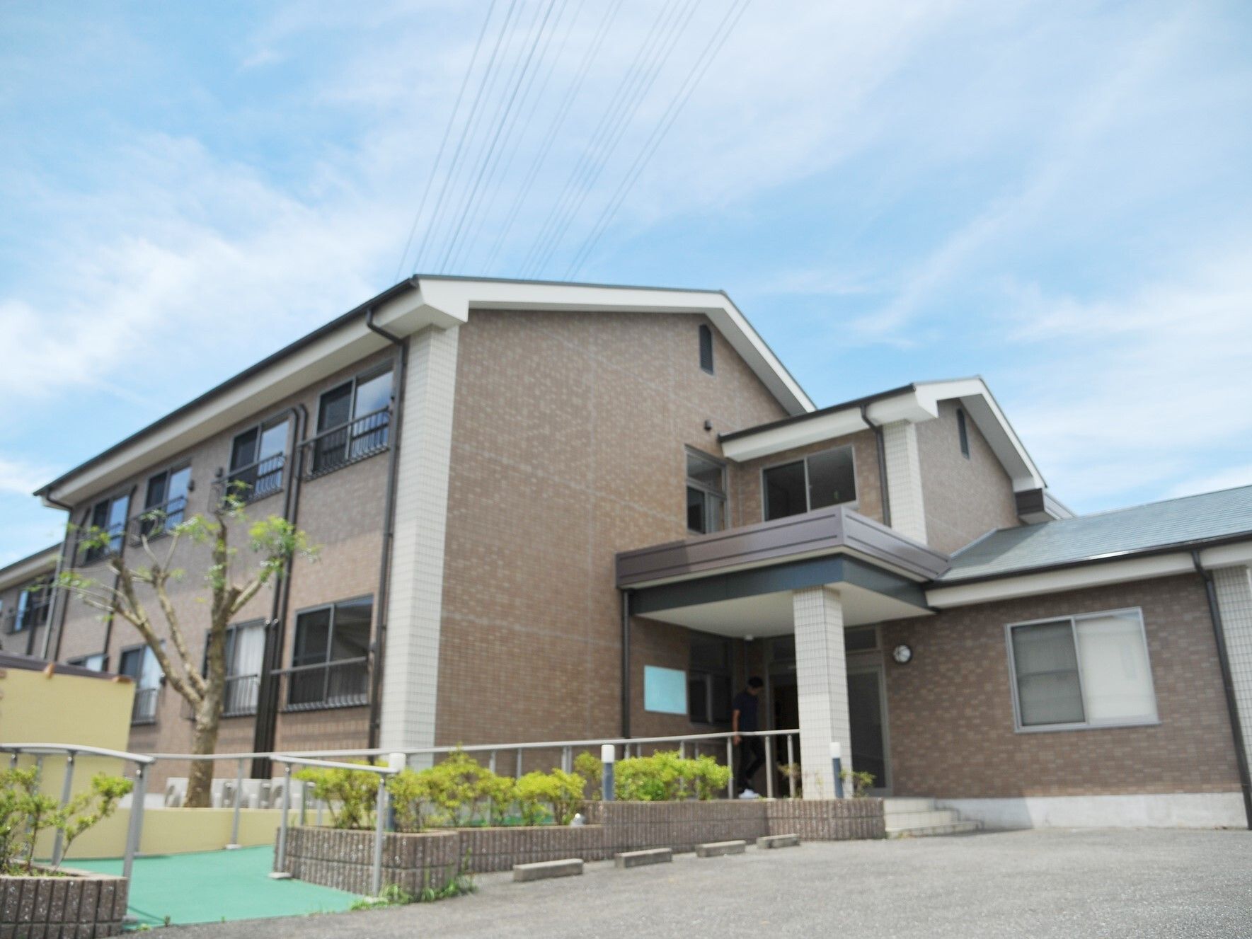 ブラウン系の2階建ての建物は三角屋根が付き、建物の玄関部分には手すり付きのスロープが設置され、敷地内には緑も植えられている。