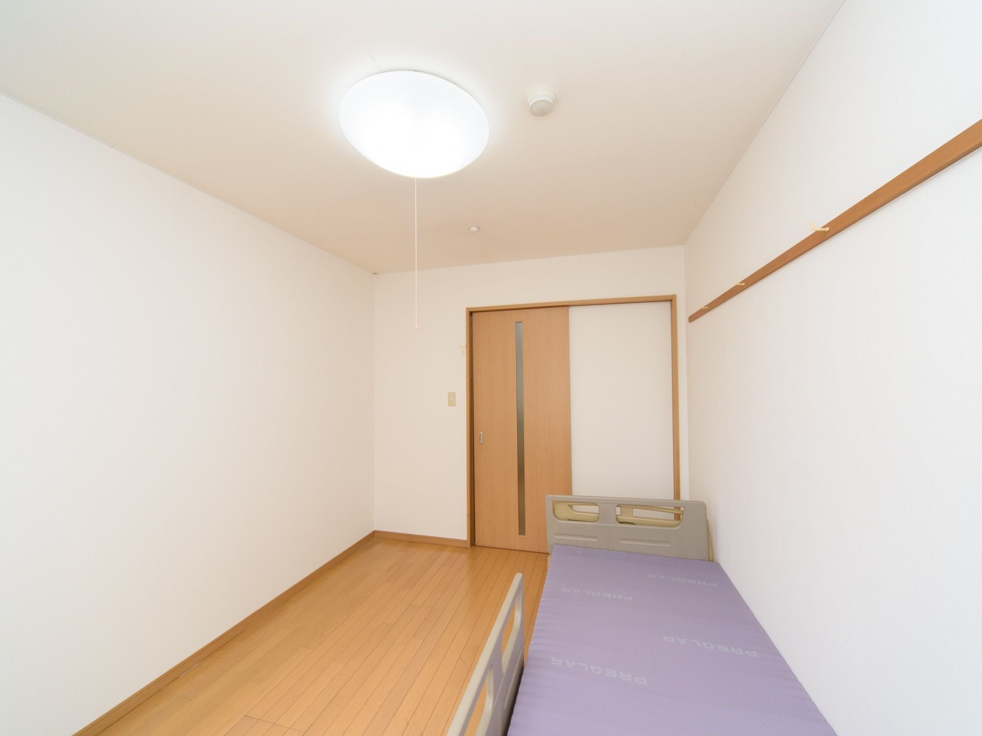 白を貴重とした居室で、フローリングの床になっている。室内にはベッドが置かれ、天井には照明器具が取り付けられている。