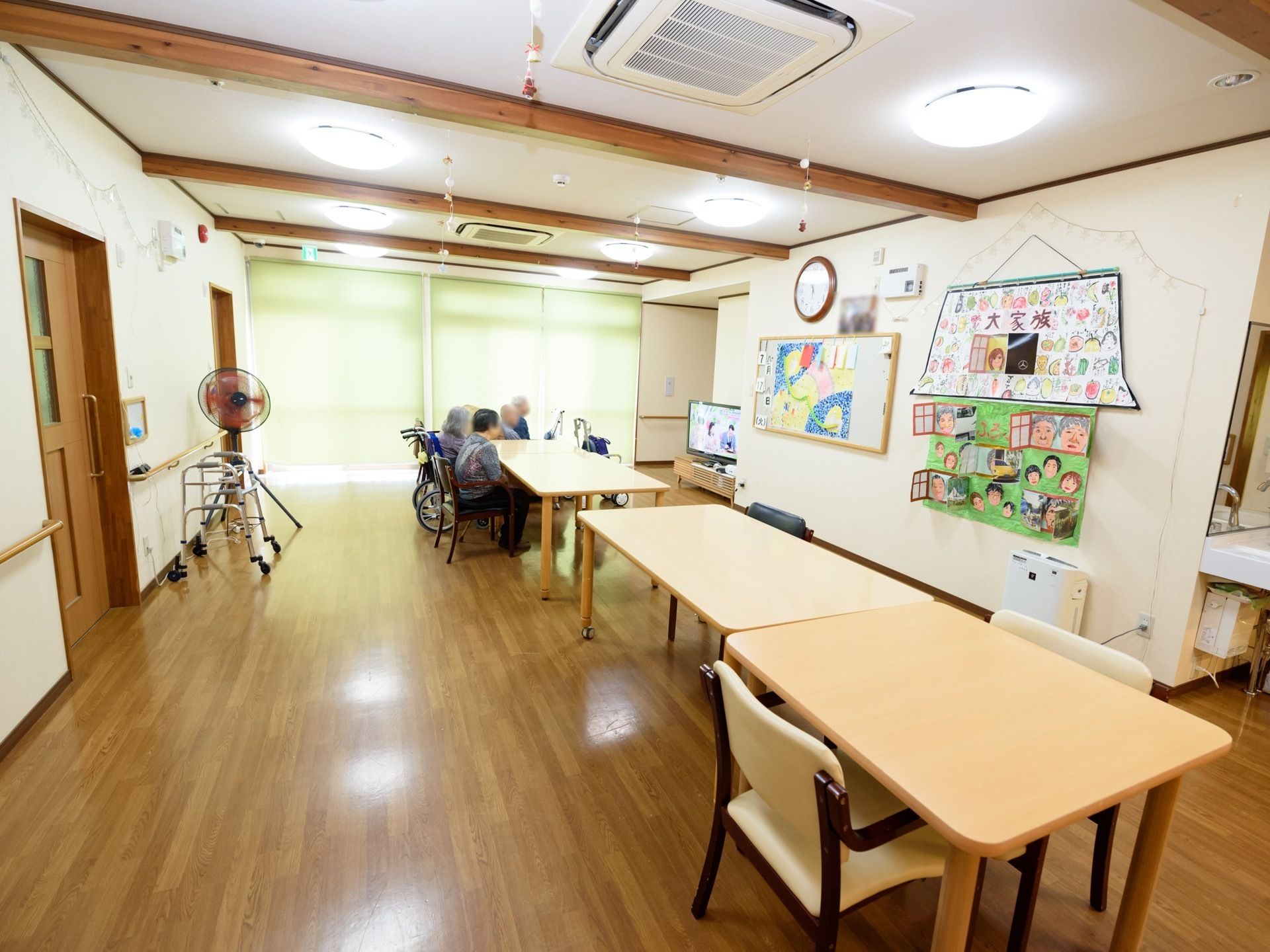 日が当たる明るい空間。テーブル数台とソファが横並びになり、入居者様がテレビを観ている。テレビ横の壁に入居者様の似顔絵などが展示されている。