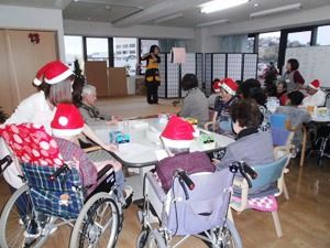 赤いサンタ帽を付けてクリスマス会に参加する入居者とスタッフ。ステージではフルート演奏が行われている。