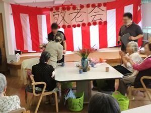 赤い垂れ幕のかかったステージ。入居者が拍手をしてお祝いしている様子。