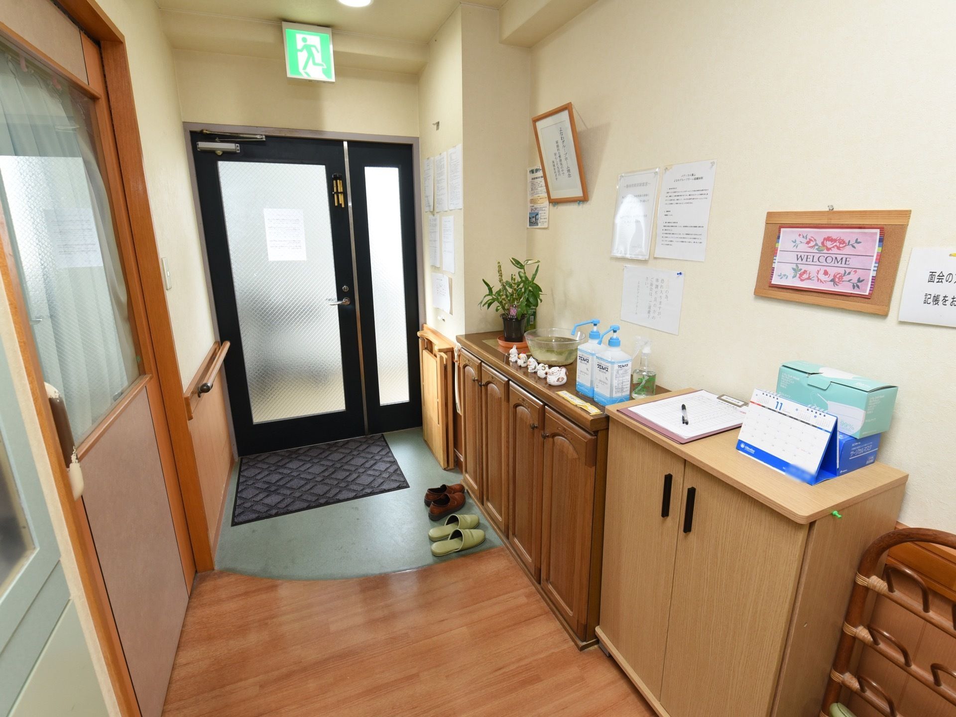 玄関扉の前に玄関マットが敷かれている。ドアの上に非常灯がある。壁には下駄箱が取りつけられ、消毒液や植木鉢が置かれている。