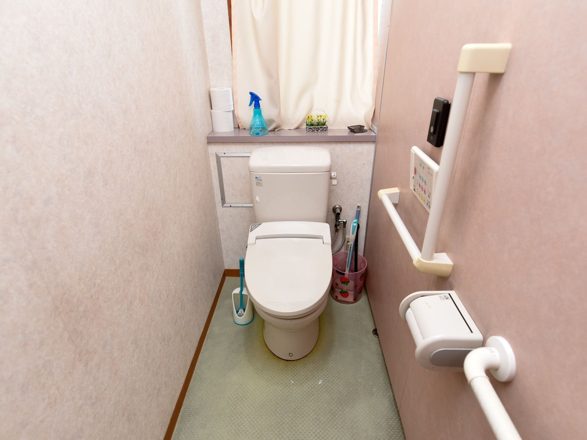 トイレは温水洗浄便座を設置しており、操作は壁のパネルで行う。床に掃除用具が置かれ、窓際に予備のトイレットペーパーがある。