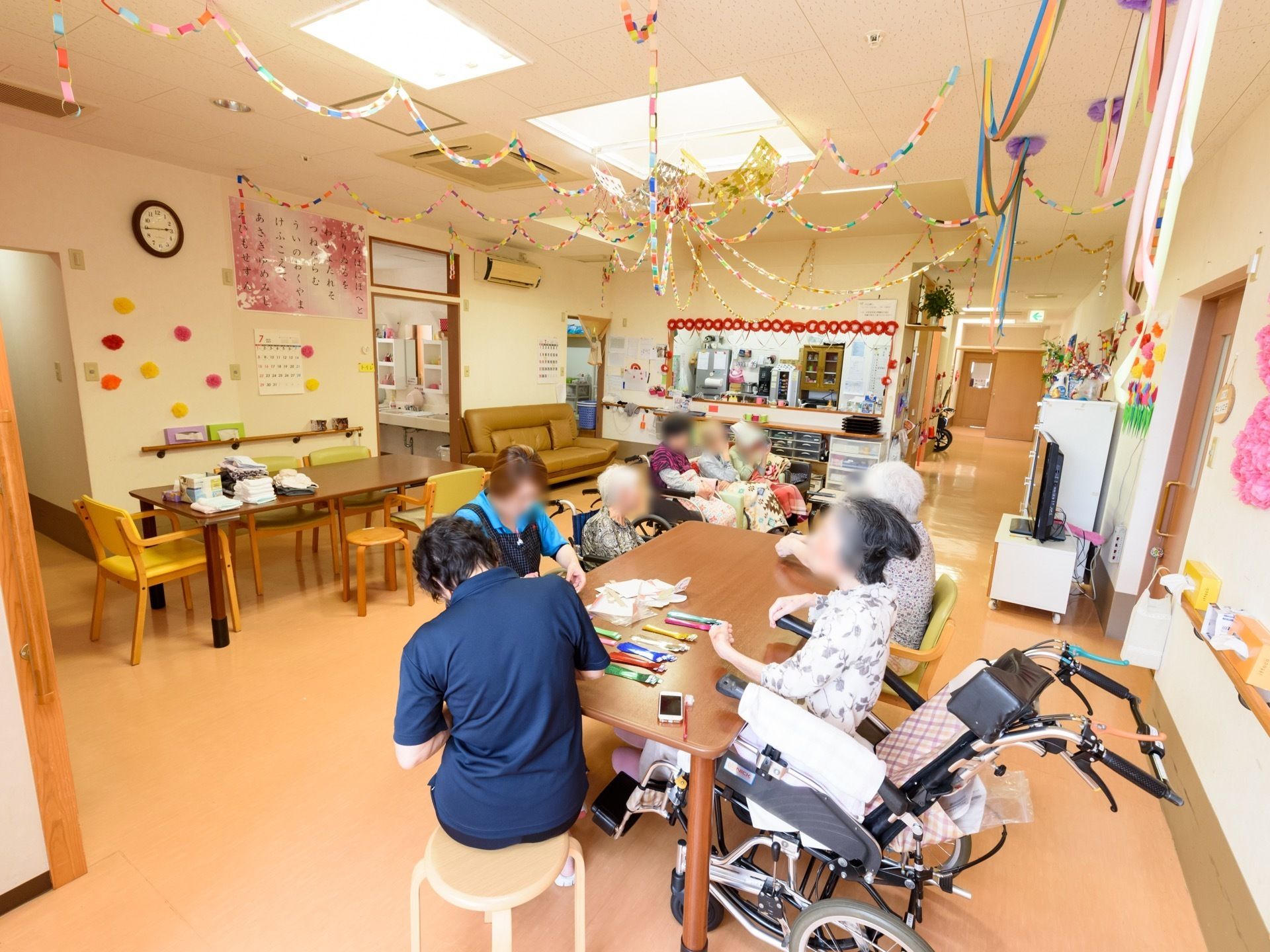 リビングのテーブルを職員と入居者様が囲んでいる。一部の入居者様は車椅子でテーブル席についているが、窮屈な印象もなくゆったりと談話している。