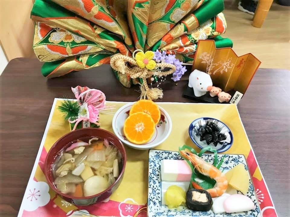 正月の干支の置物や金屏風の前には雑煮やおせち風のごちそうが並んでいる。