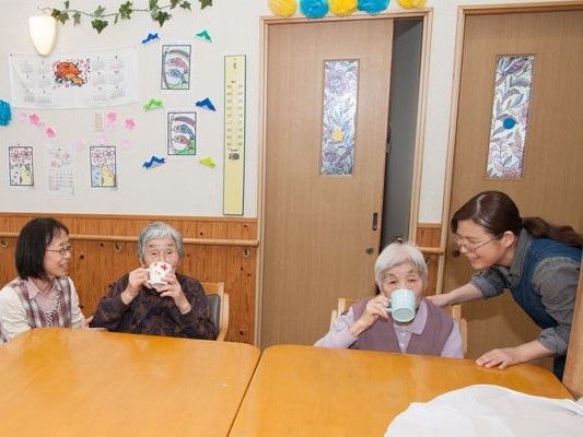 2名の女性入居者がコップで飲み物を飲んでいて、そのそばでそれぞれスタッフが見守っている様子。
