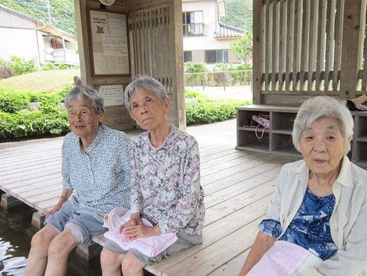 広いウッドデッキに荷物置き場が付いた屋外の足湯で足を温めている入居者3名の様子。
