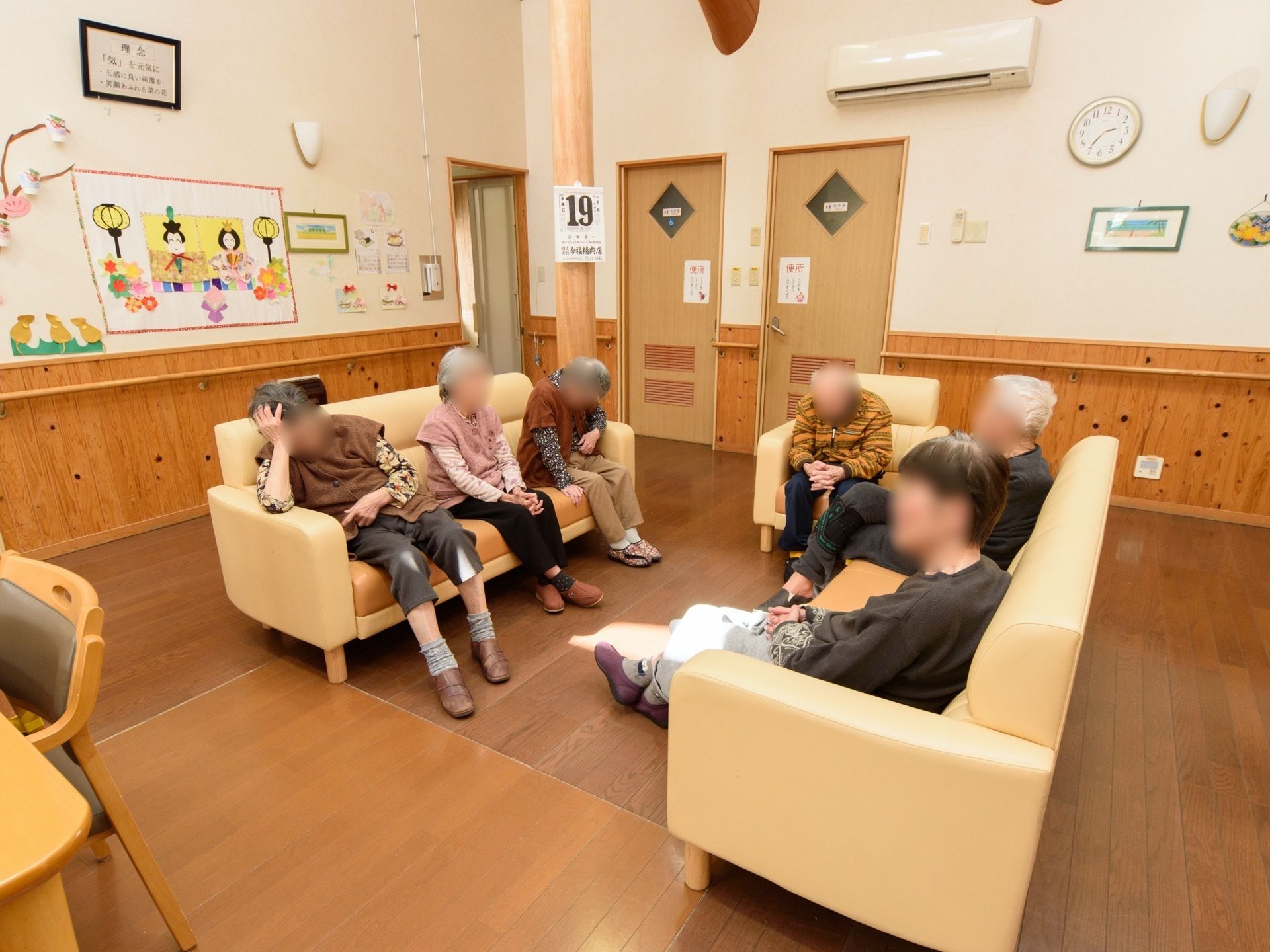 柱に日めくりカレンダーが掛けられ、壁に季節のレイアウトが施された談話スペースである。ソファーに腰掛けられている数名の入居者様の姿が見える。
