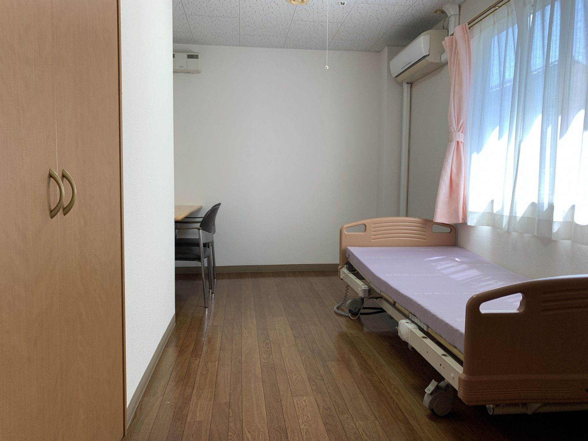 黒いカーディガンを着た車いすの高齢女性の向かいには髪の長い女性がいて、女性が差し出す手を見て高齢女性が笑っている。