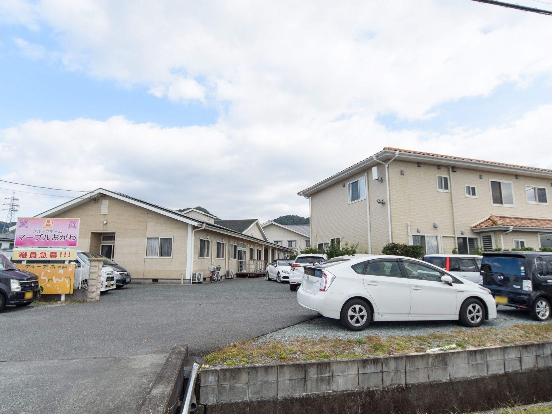 ピンクを基調とした、平屋建ての施設。駐車スペースが確保され、車が停まっている。周囲には住宅が建っている。