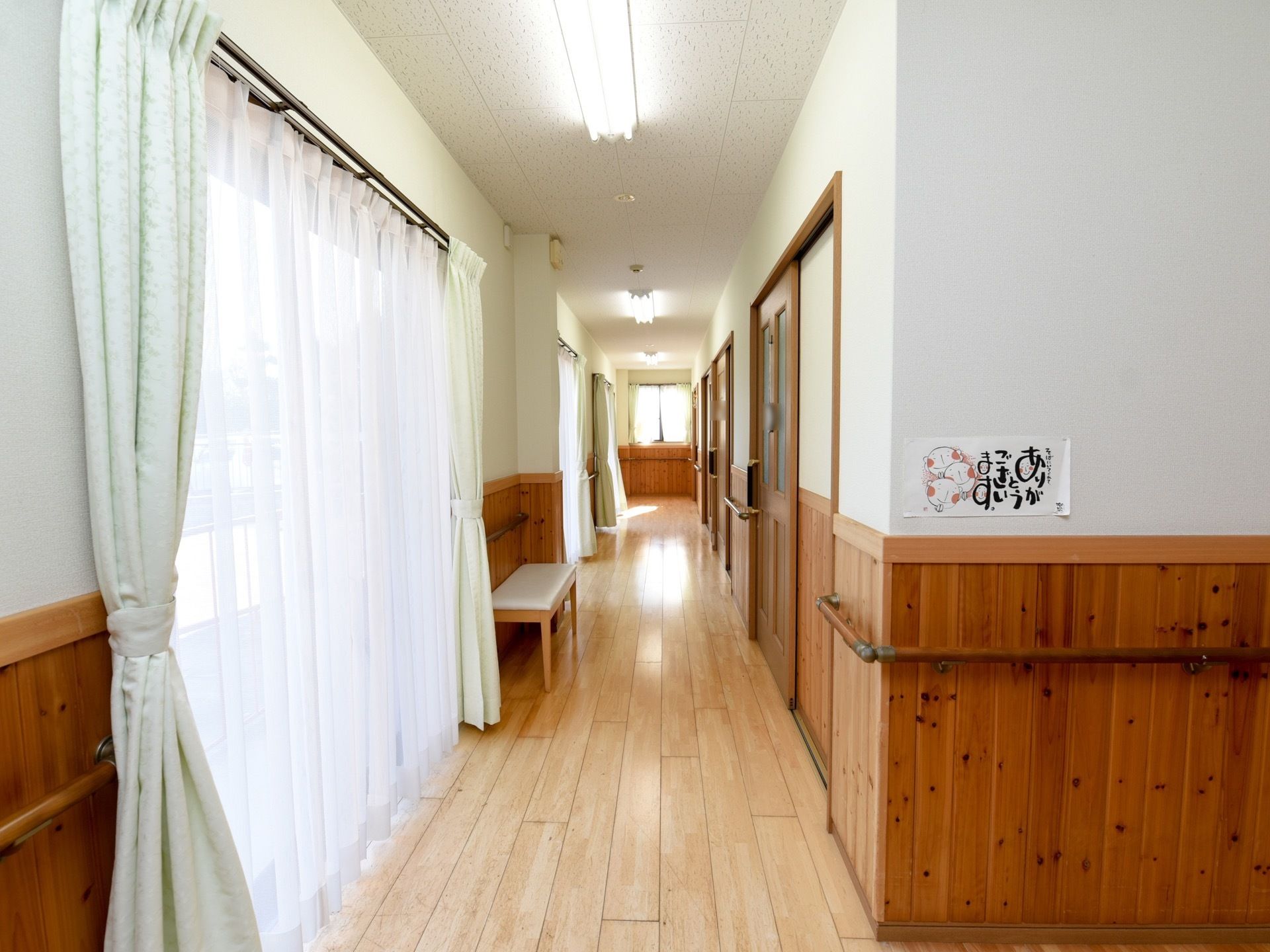 廊下の左側に窓が並び、薄手のカーテンが引かれている。窓の間にベンチが置かれている。右側は居室へのドアが並んでいる。