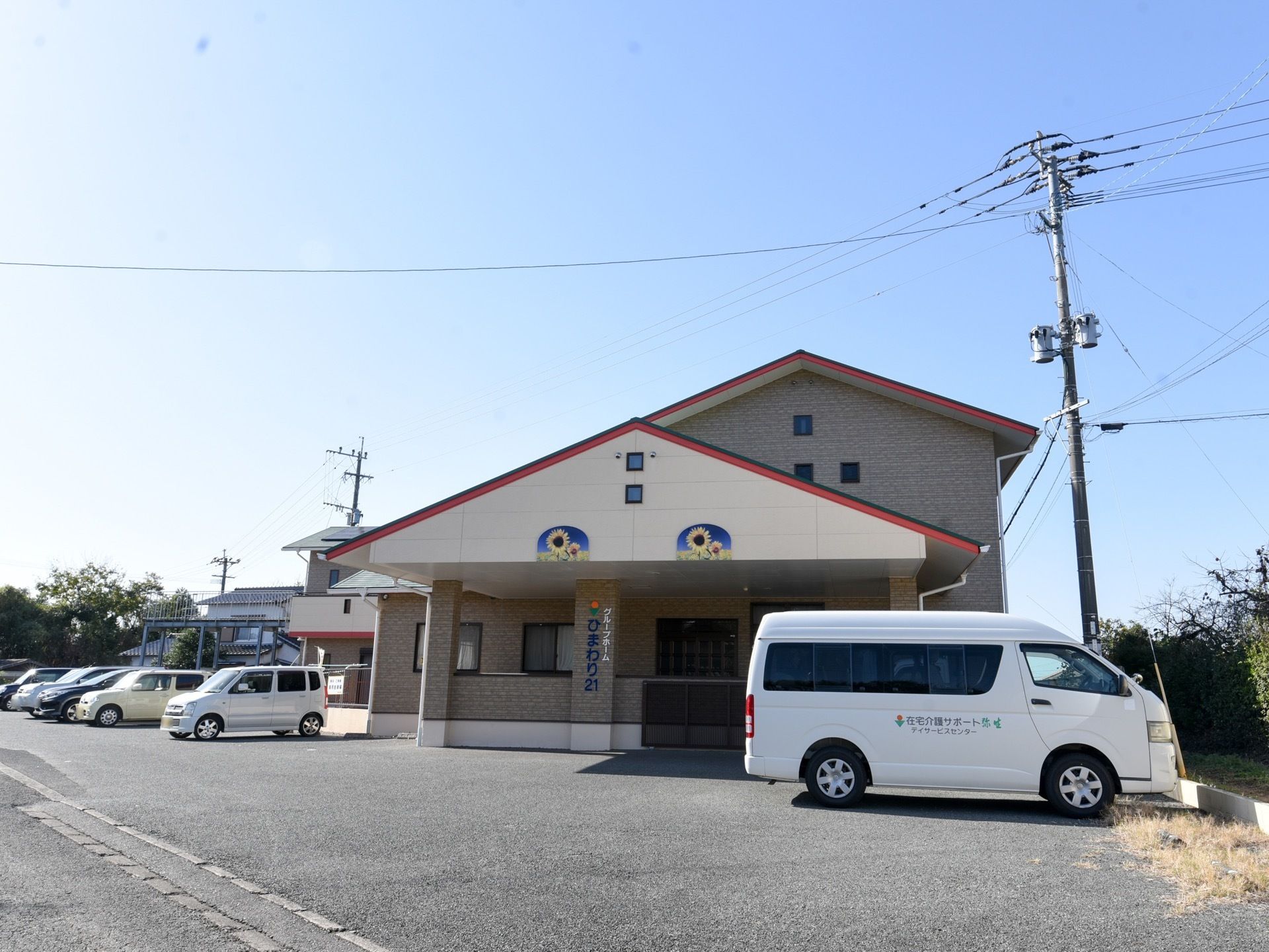 レンガのタイルが貼られた居住部と、白い壁の屋根裏のついた建物に、赤い三角屋根が乗っている。入口前は駐車場になっている。
