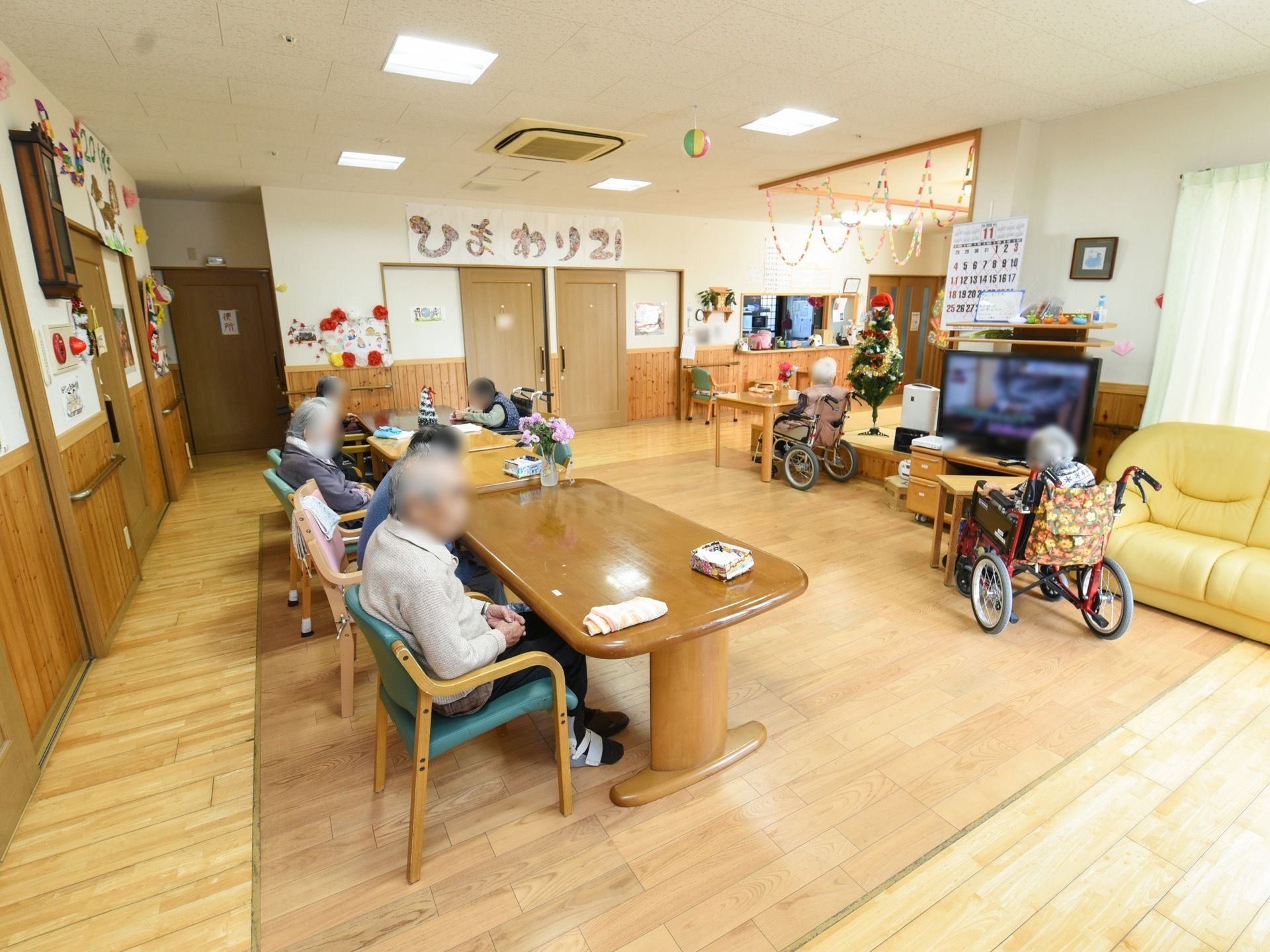 ４人掛けのテーブルセットが横に３台並べられている。椅子にはたくさんの入居者様が座っている。テーブルに花が飾られている。