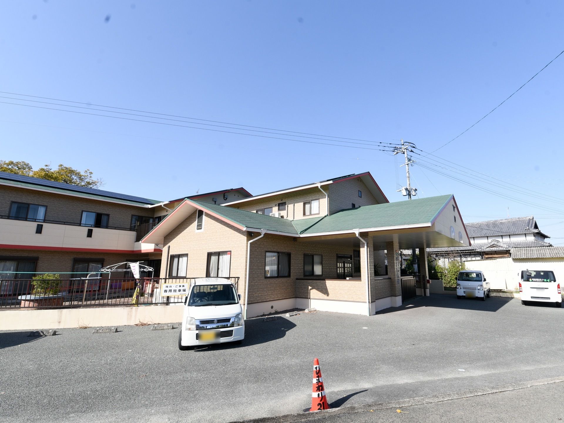 ベージュの壁の２階建ての建物で、三角の屋根が乗っている。建物の前は広い駐車スペースがある。敷地内に中庭がある。