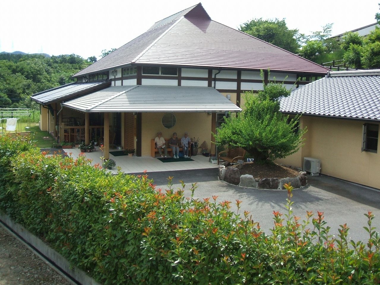 古民家風の建物の周りには自然が多く、生垣もある。玄関のベンチには数名の入居者が座っている。