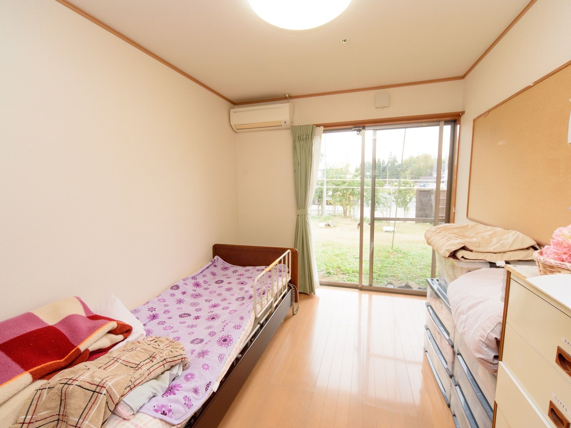 大きな窓があり、日当たりと風通しが良い居室。窓からは緑豊かな自然が見える。柵付きの介護用ベッドと、衣装ケースなどが置かれている。