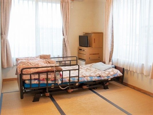 寝具付きのベッドが置かれた和室。大きな窓が二つ付き、テレビがセットされたラックがある。