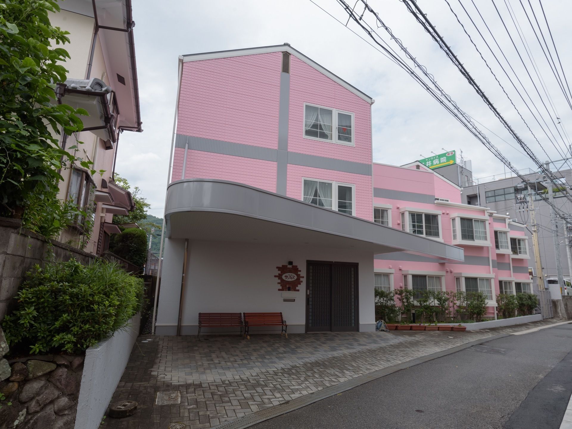 パステルピンクの外壁が特徴的な３階建ての施設である。建物に沿って庭木が植えられている。大きめのひさしがエントランスに設置されている。