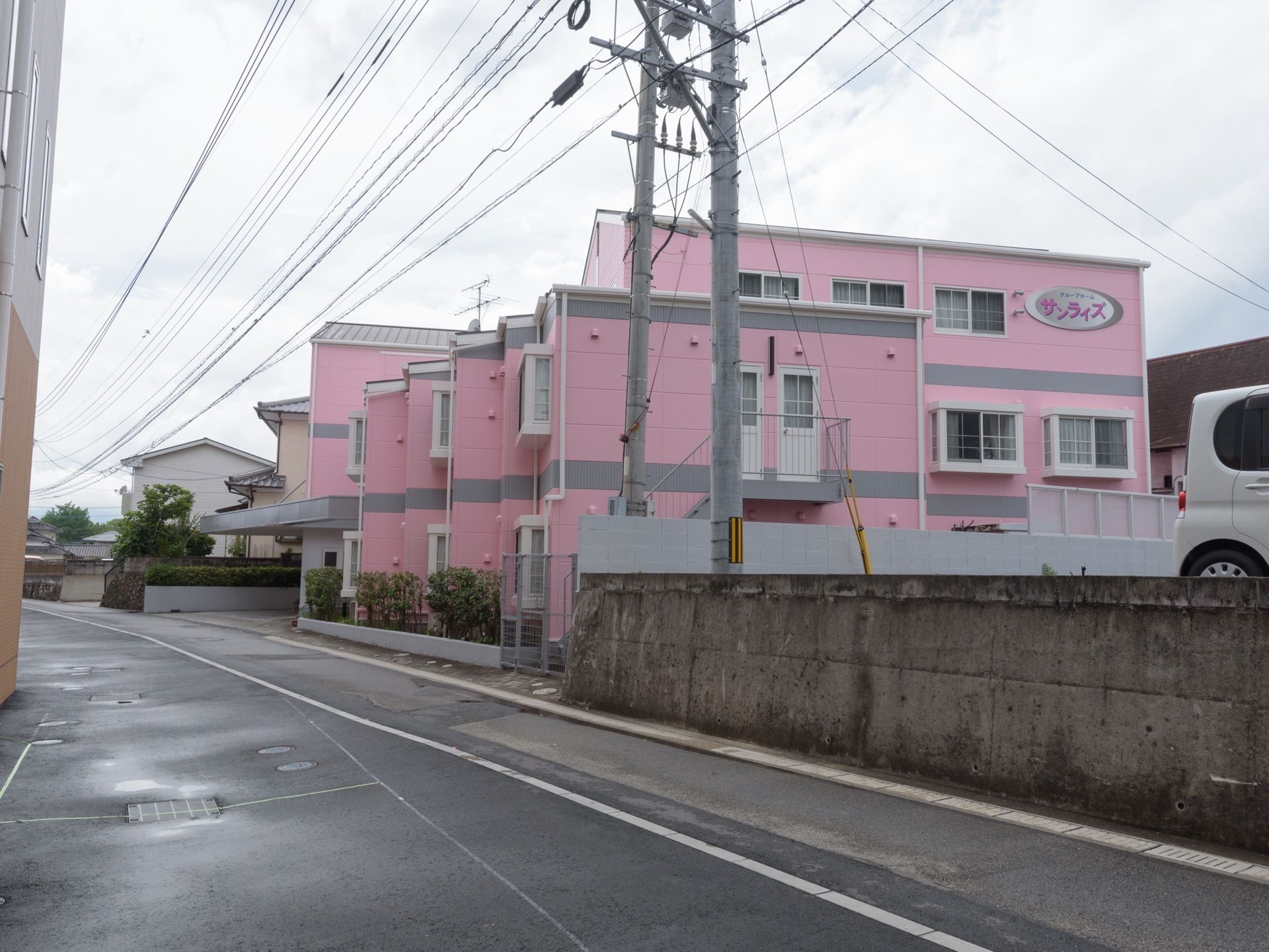 施設はパステルピンクにグレーのラインが入った３階建ての建物である。外壁には、施設名が記された楕円形の看板が設置されている。