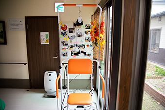 車いすの高齢男性と付き添いの若い女性スタッフが緑豊かな遊歩道で何かを眺めている。