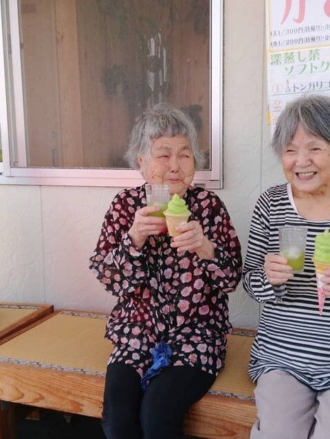 白い服を着た高齢女性と青いユニフォームを着た男性スタッフが並んで体操をしている。