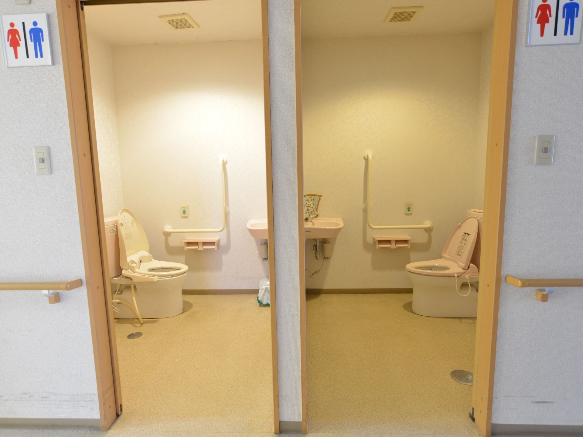 2つの男女兼用トイレが線対称に並んでいる。タンク付きのトイレで便座の横には、手すりがつけられている。